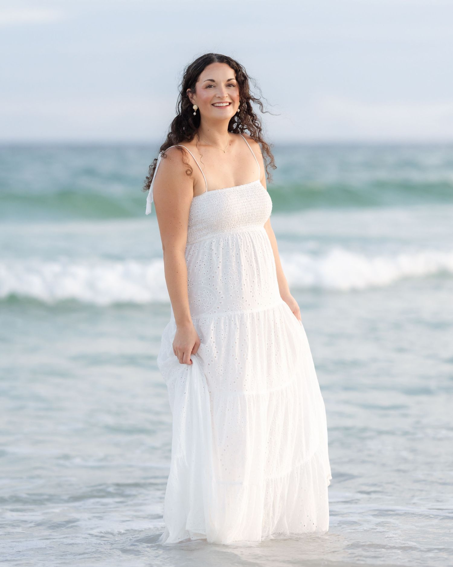 Smiling woman in white sundress standing in ocean waves on beach.