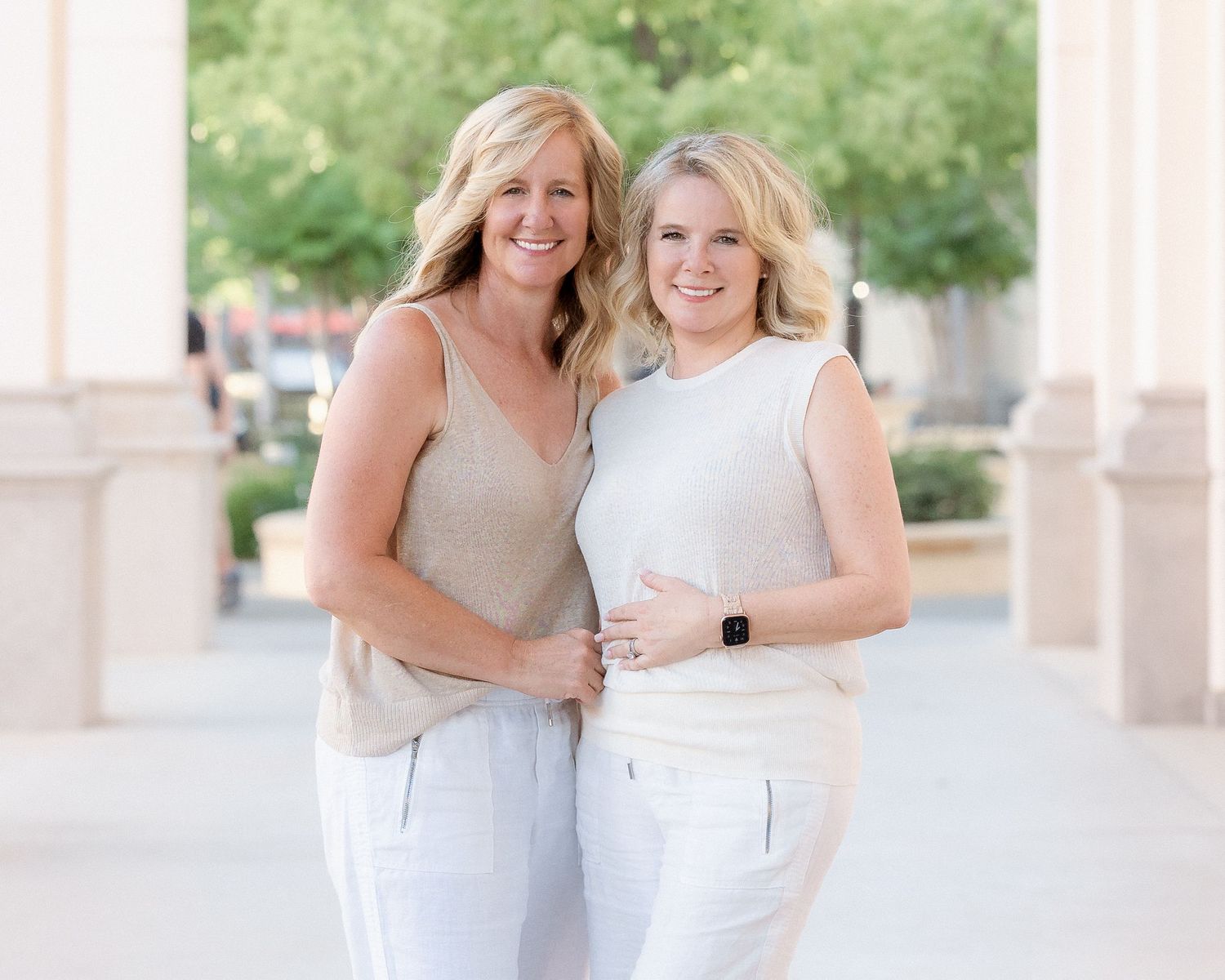 Two smiling blonde women in matching neutral outfits posing together outdoors near elegant columns.