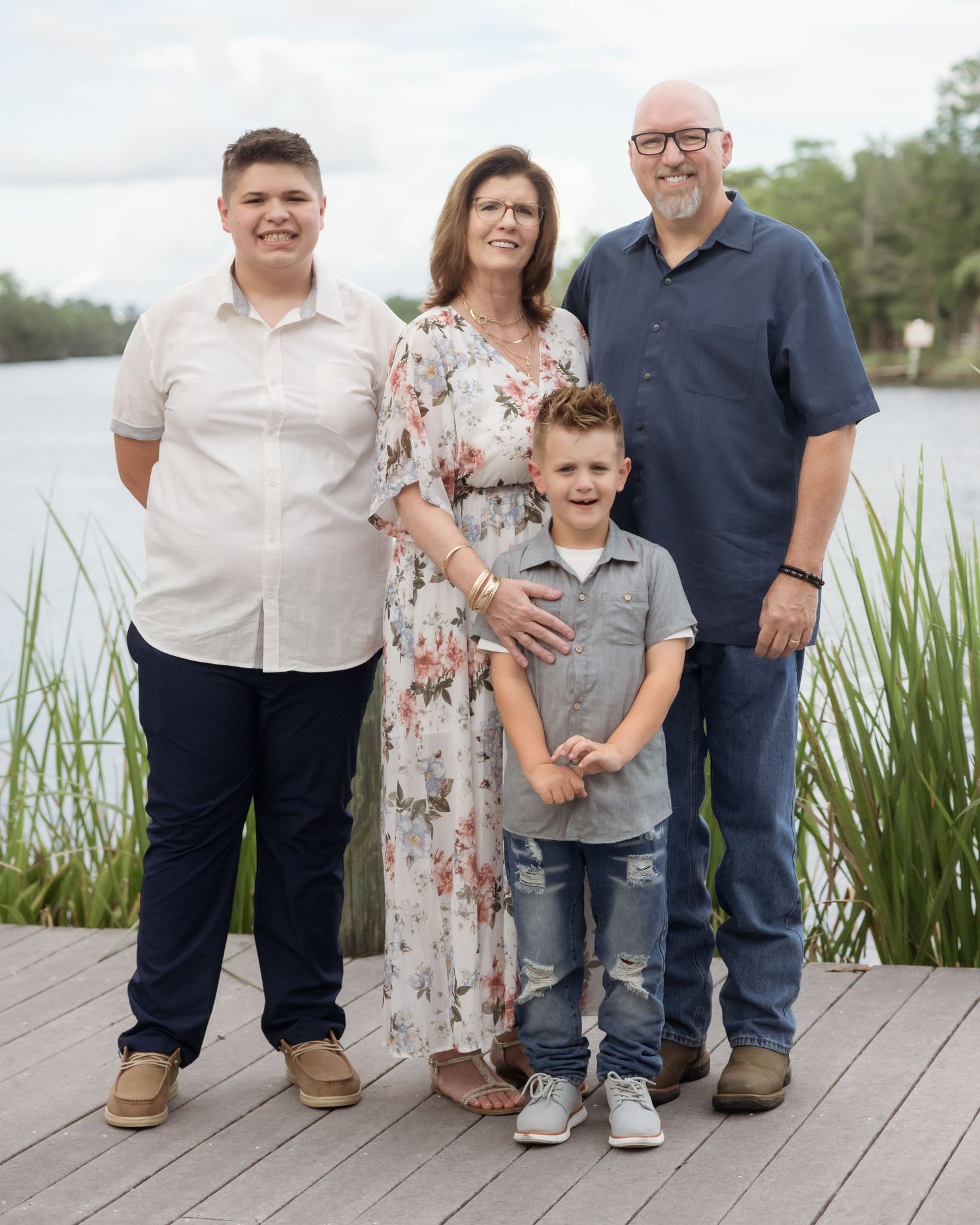 Family of four posing on a wooden dock by a lake, with tall grass and water in the background.