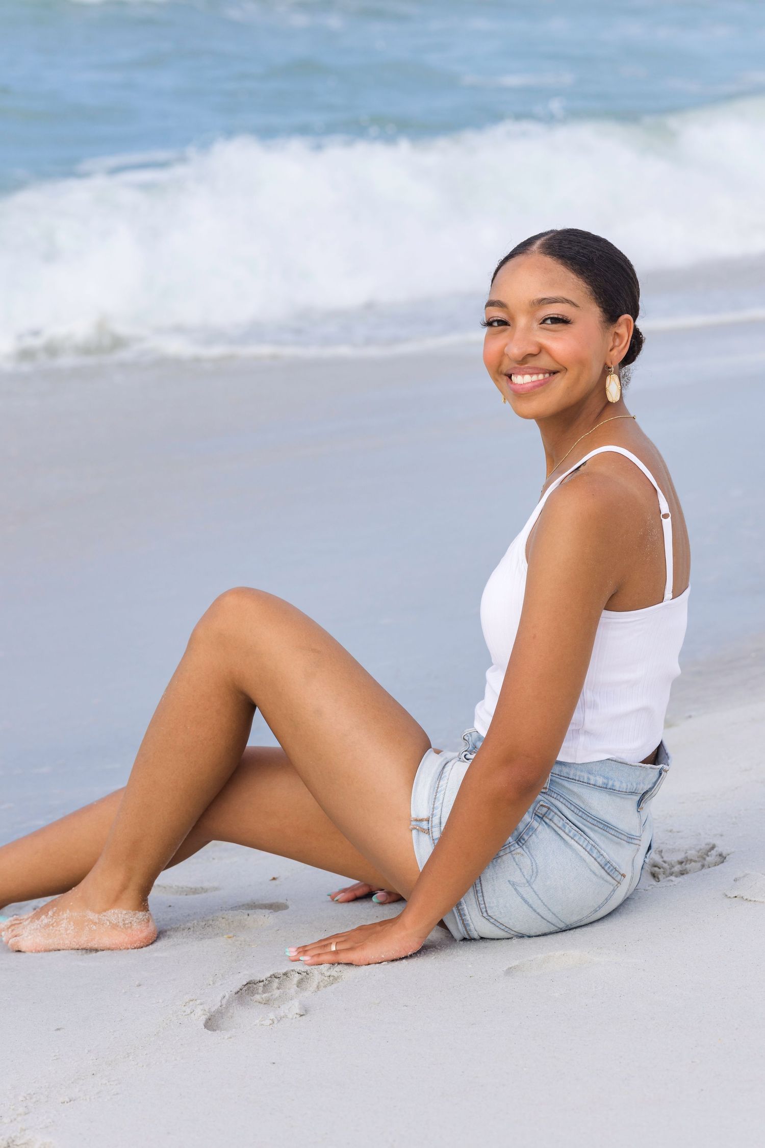 Young woman in white tank top and denim shorts smiling while sitting on sandy beach with ocean waves behind her.