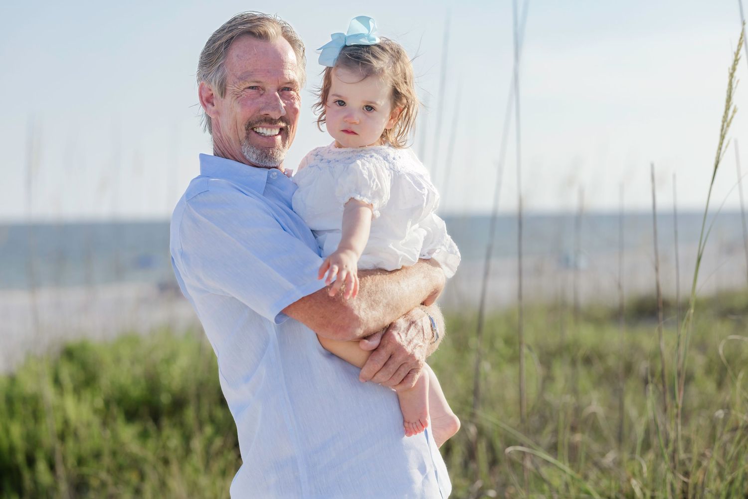 Man in blue shirt smiling and holding a toddler girl with blue bow at beach.