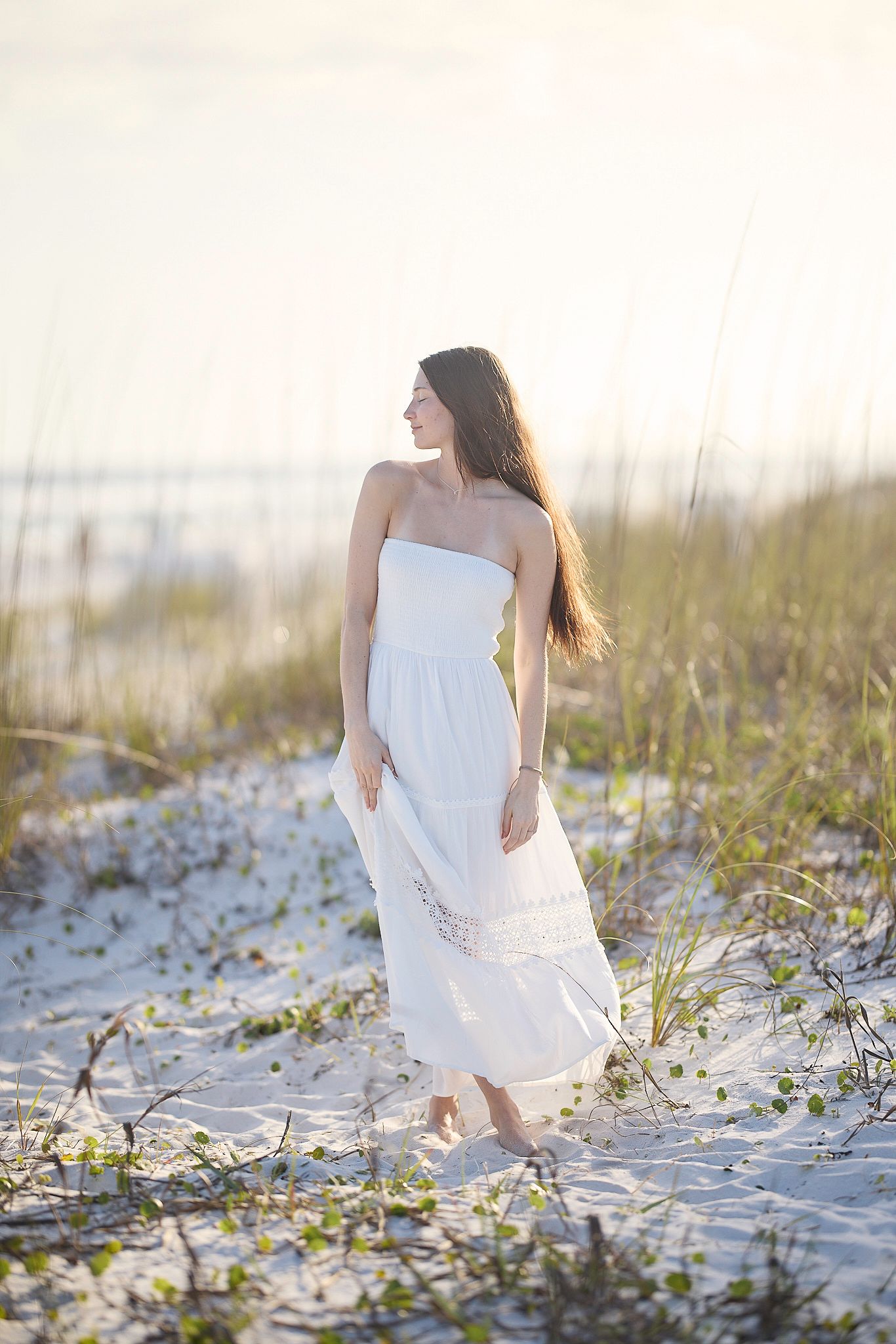 Young woman in flowing white strapless dress standing among sand dunes and sea grass at golden hour beach.