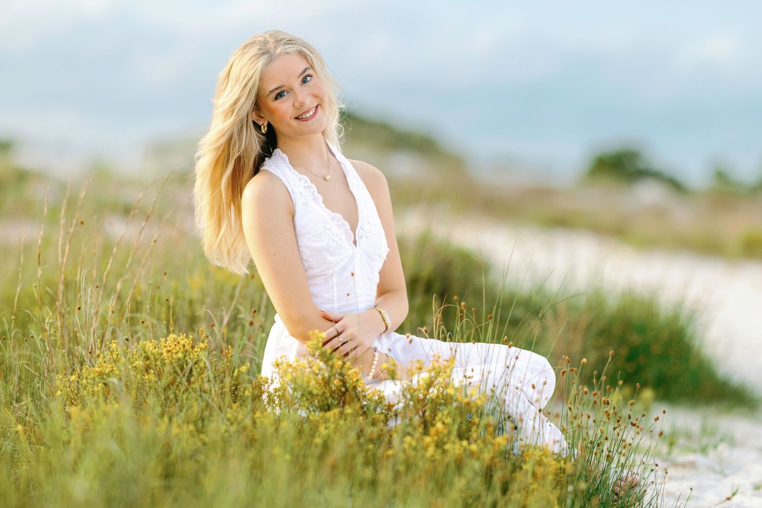 Smiling blonde woman in white outfit sitting in a sunny meadow with wildflowers on a beautiful day.