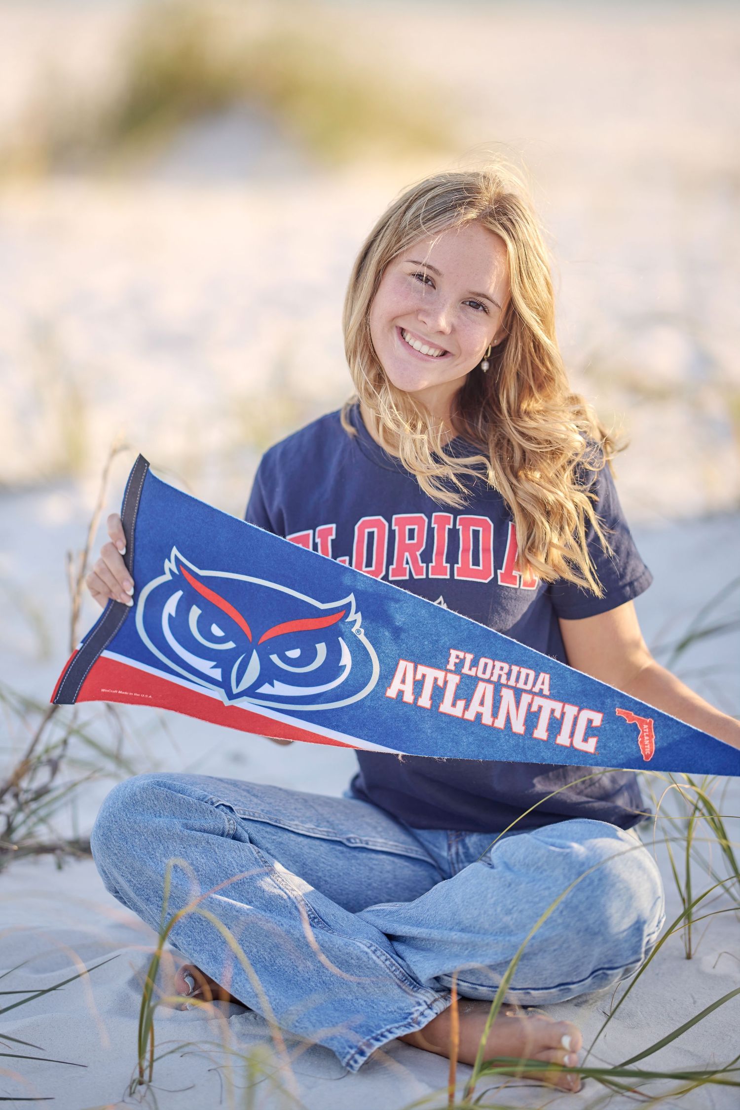 Smiling blonde woman sitting on beach sand holding a Florida Atlantic University pennant wearing FAU shirt and jeans.