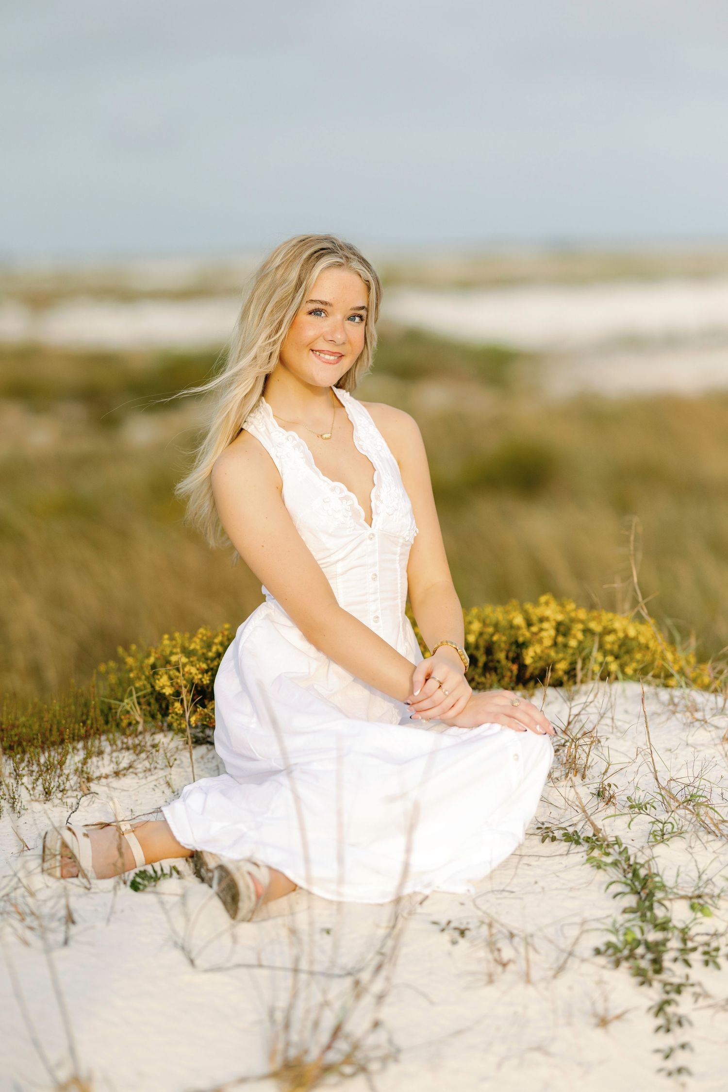 Person in white lace dress poses serenely among yellow wildflowers on coastal sand dunes.