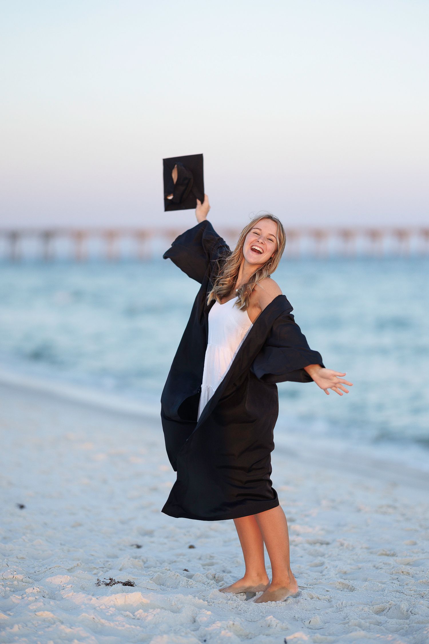 Graduate in black blazer celebrates on sandy beach at sunset with diploma in hand.