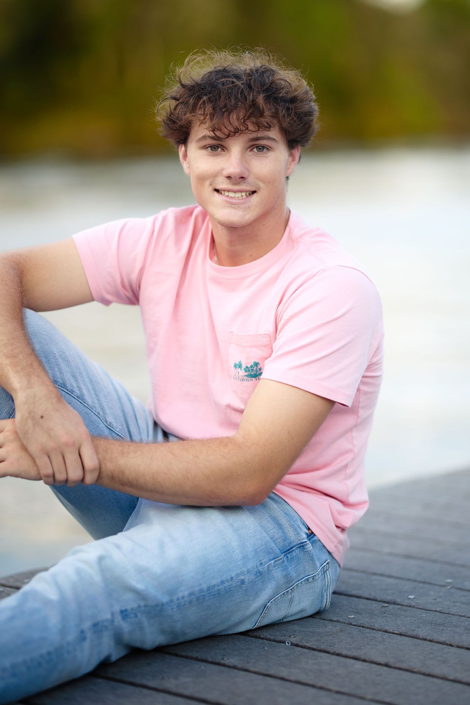 Young man in pink t-shirt and jeans sitting on a wooden dock, smiling outdoors near water.