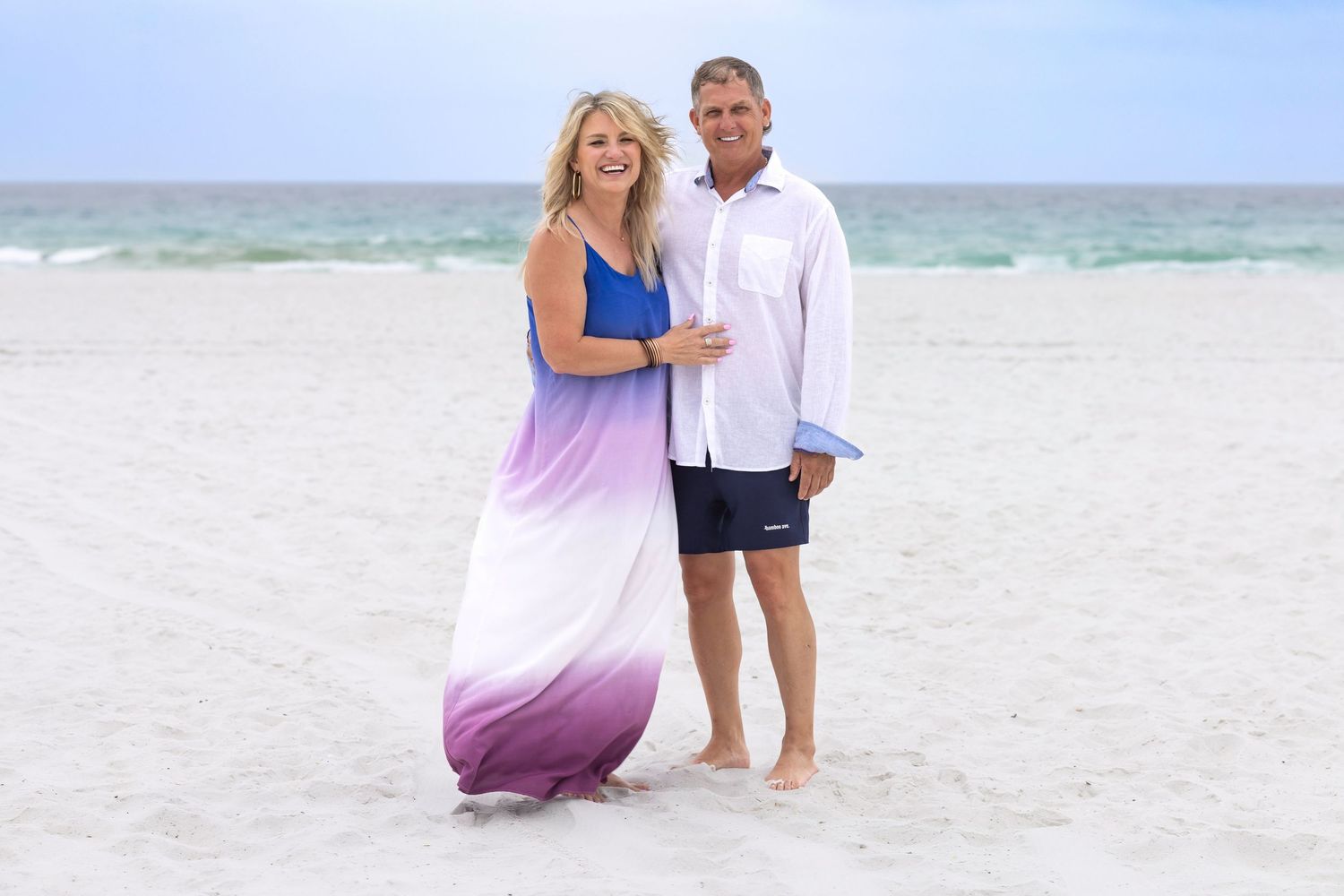 Couple posing on a white sandy beach, woman in purple ombre maxi dress, man in white shirt and navy shorts.
