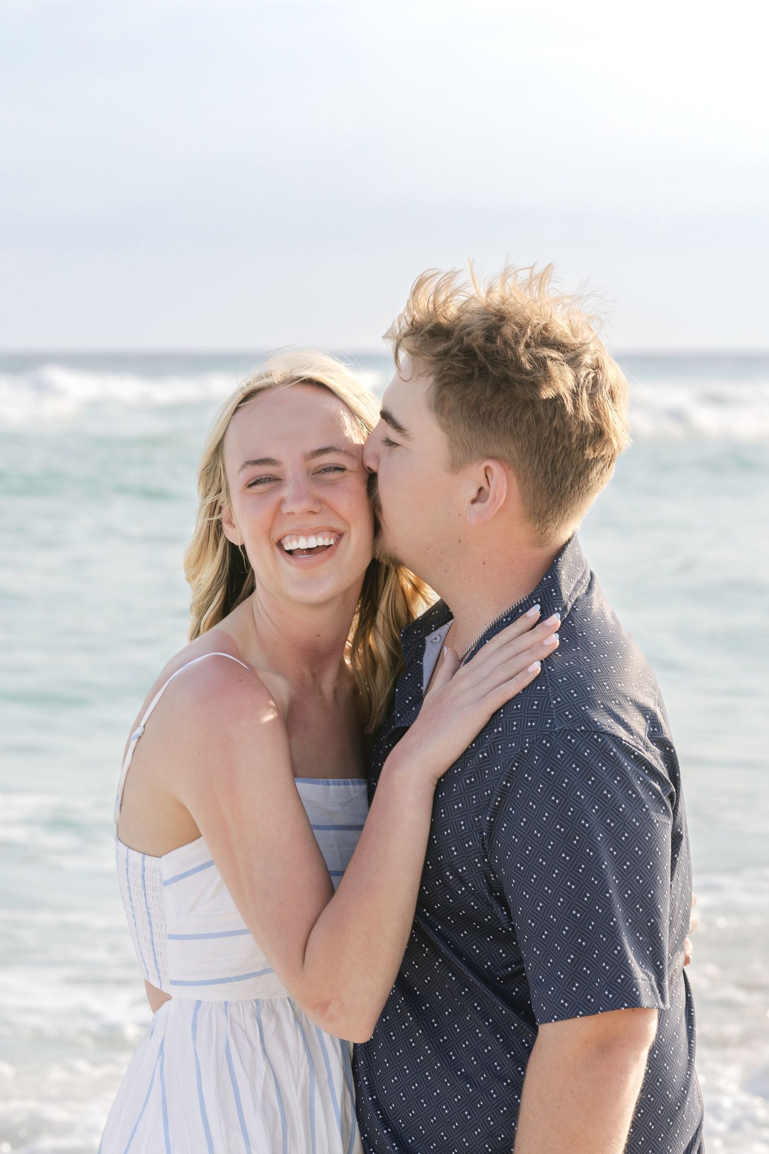 A happy couple shares a romantic kiss on the beach, with ocean waves in the background.