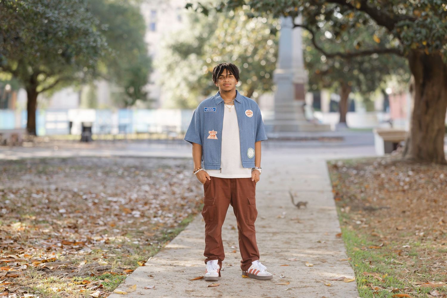 Young man in denim shirt and brown pants standing on a park pathway surrounded by trees.