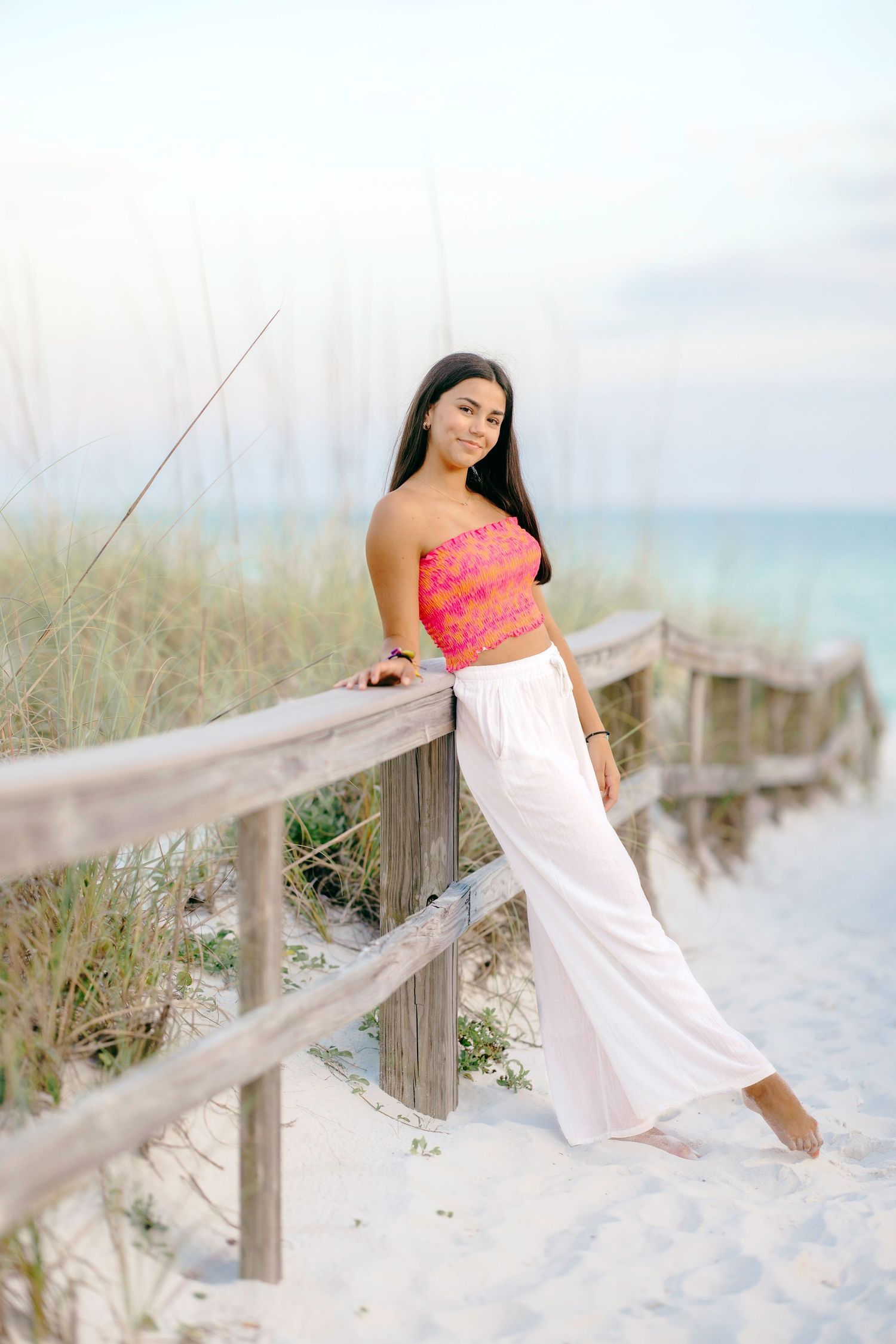 Person wearing coral top and white dress poses gracefully against beach boardwalk with ocean view.