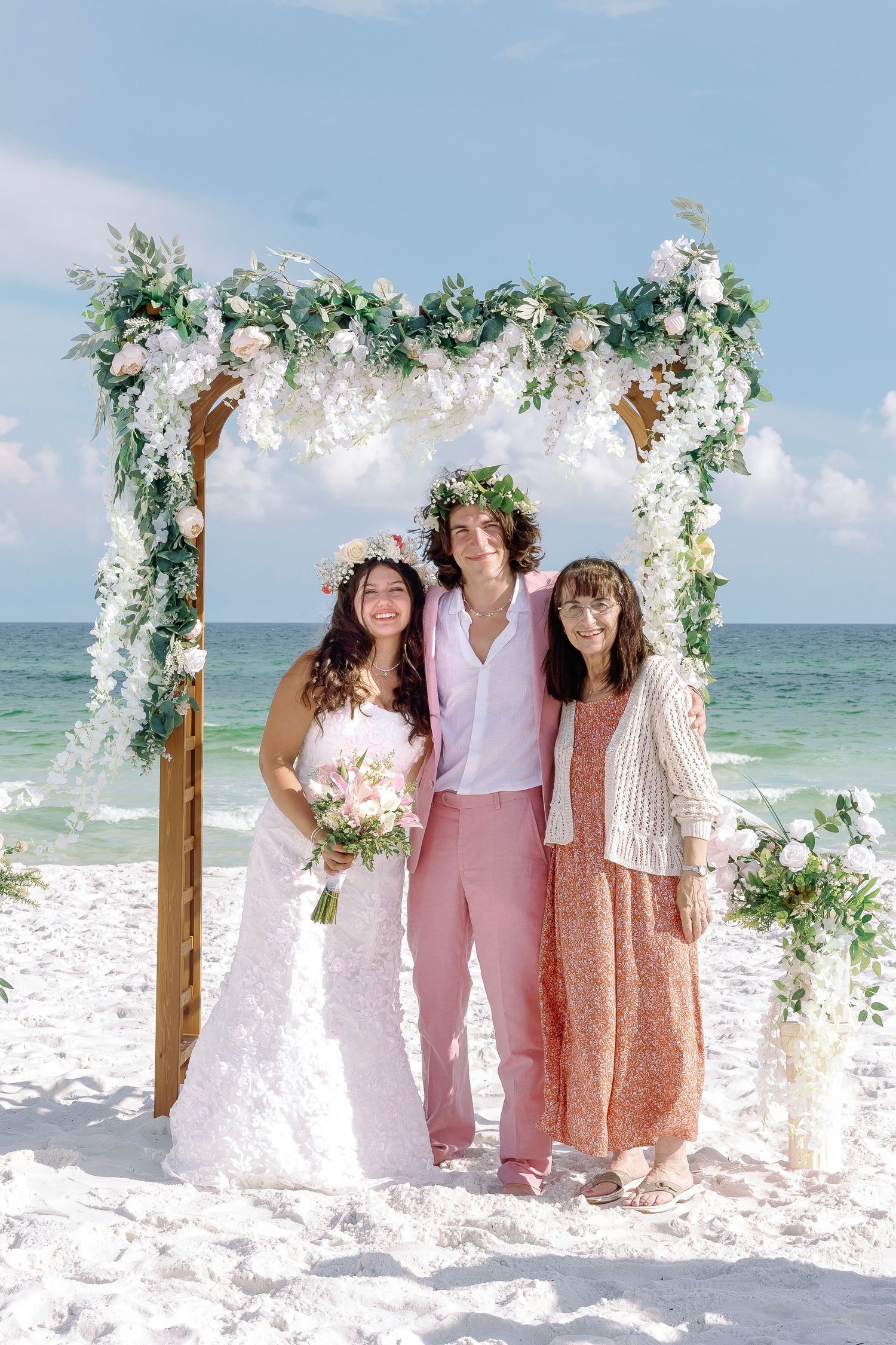 Three people stand under ornate floral arch at beachside wedding.