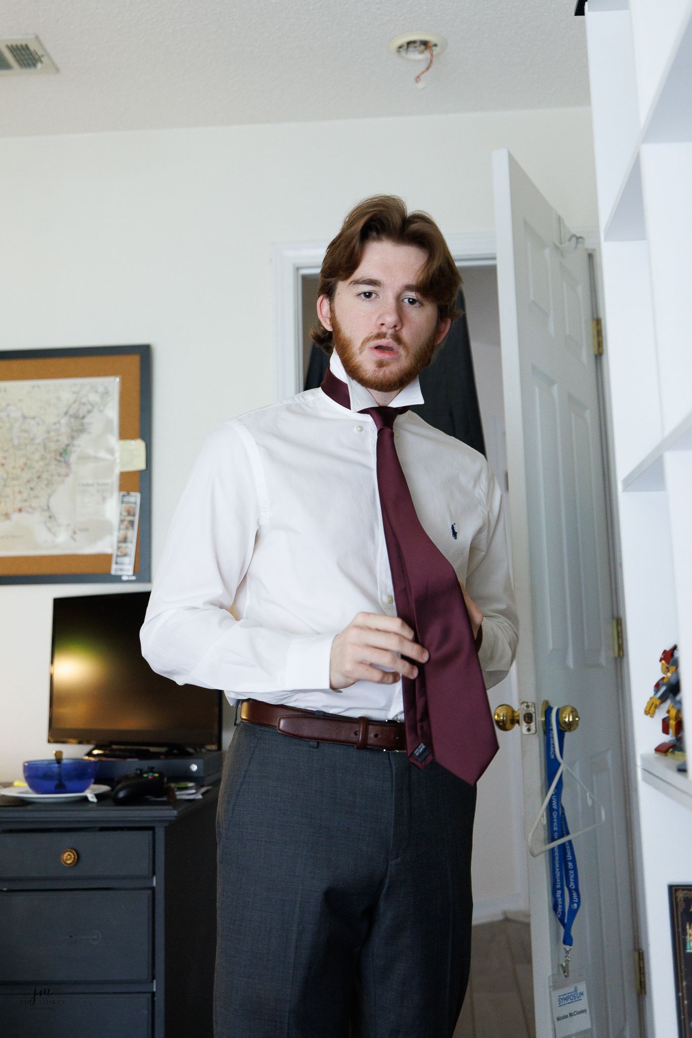 A man adjusts his burgundy tie while wearing a white dress shirt and grey dress pants.