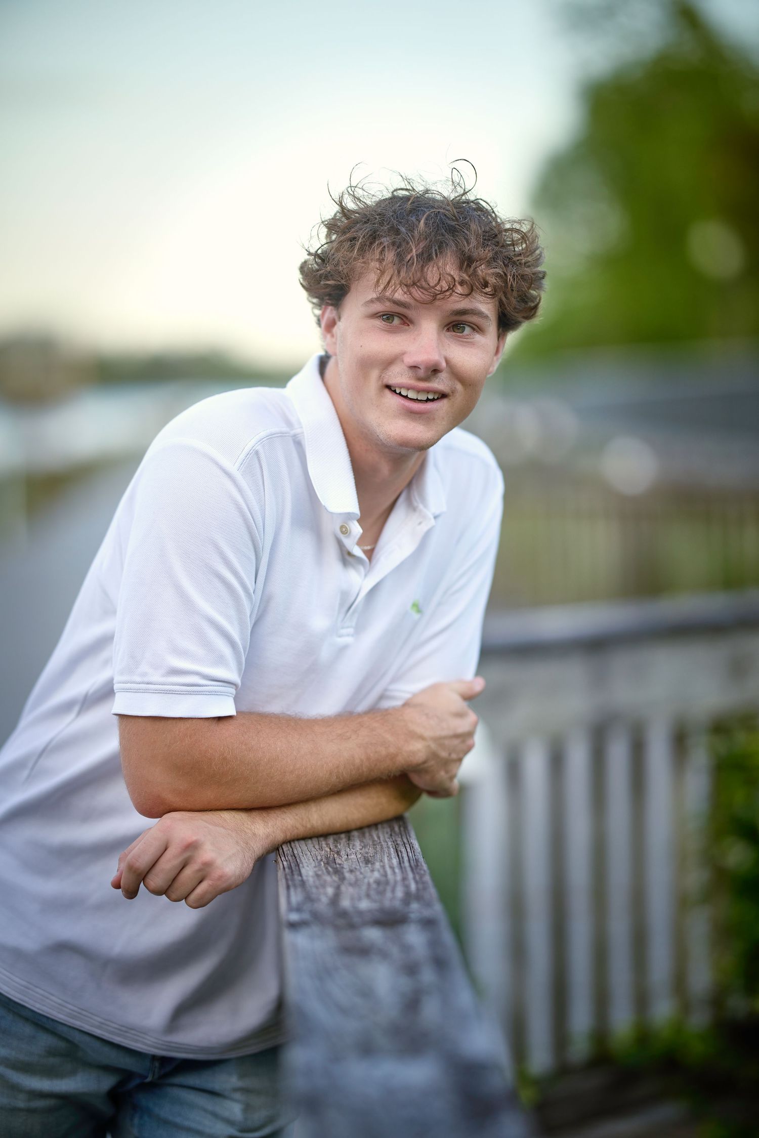 A person in a white polo shirt sitting outdoors smiling and leaning forward against a railing.