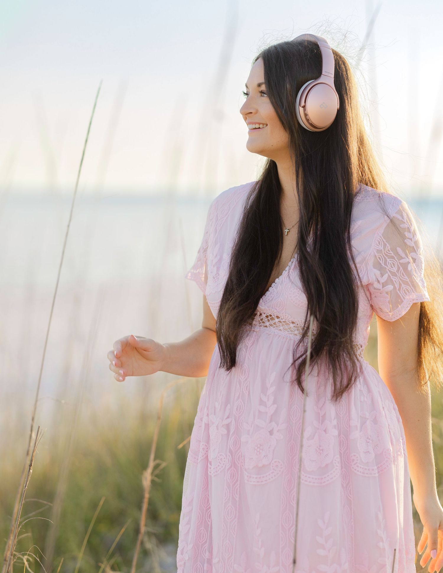 Woman in pink dress wearing headphones stands peacefully in sunlit meadow at golden hour.