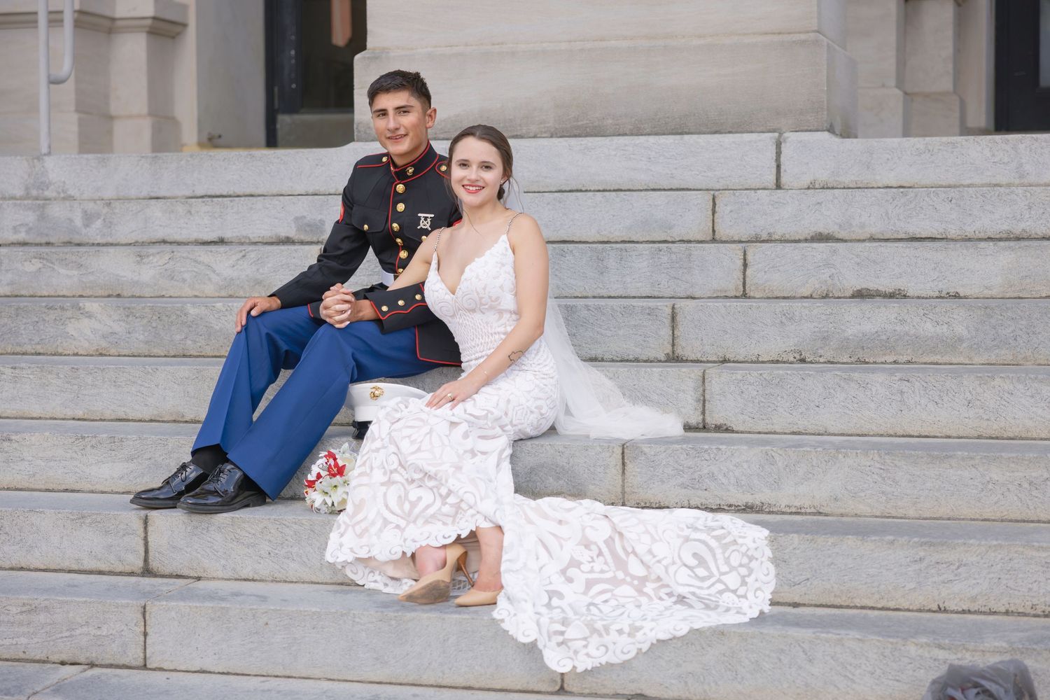 Marine in dress blues sits with bride in lace wedding gown on stone steps, smiling together on their wedding day.