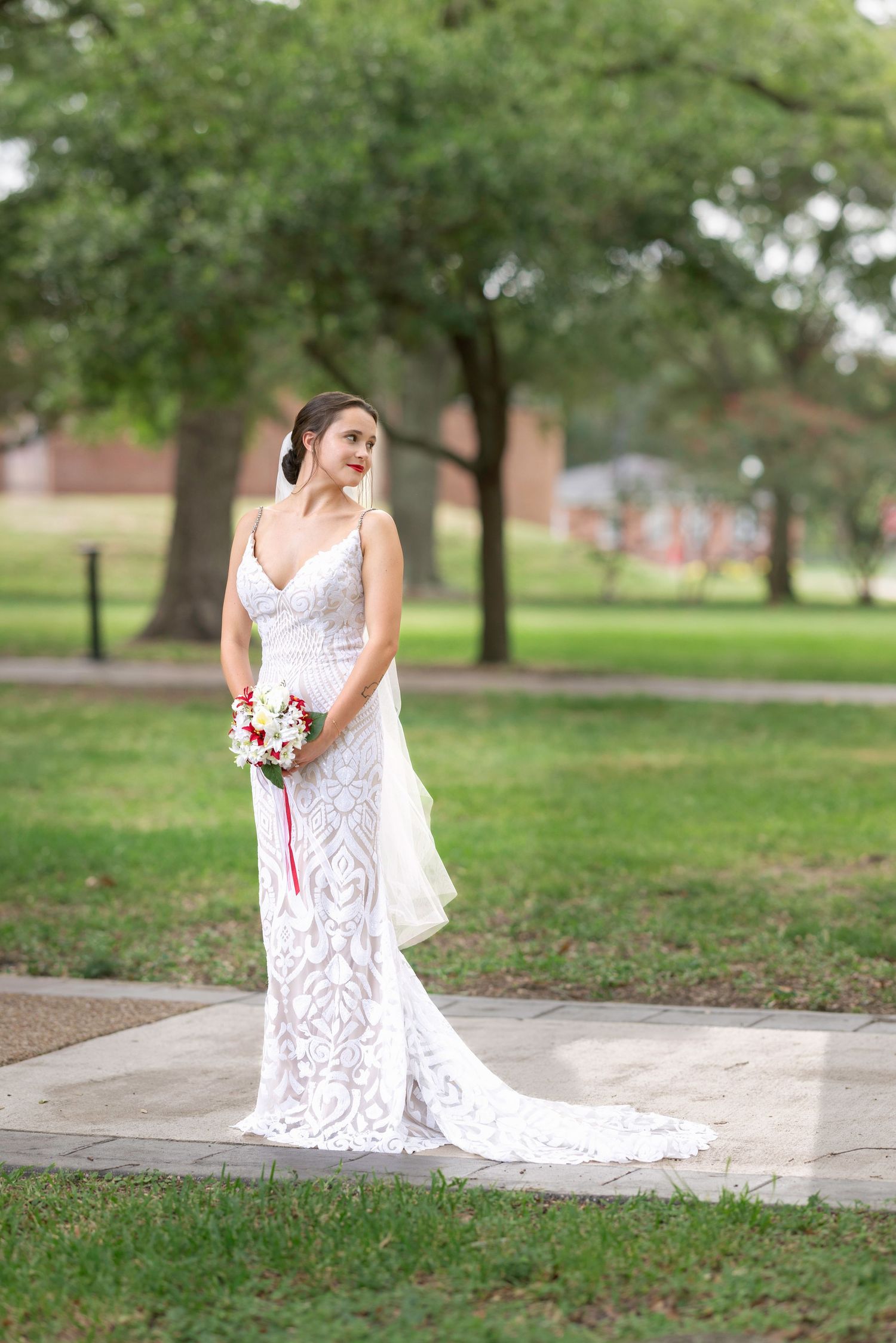 Bride in white lace gown holding floral bouquet standing on stone path in lush green park setting.