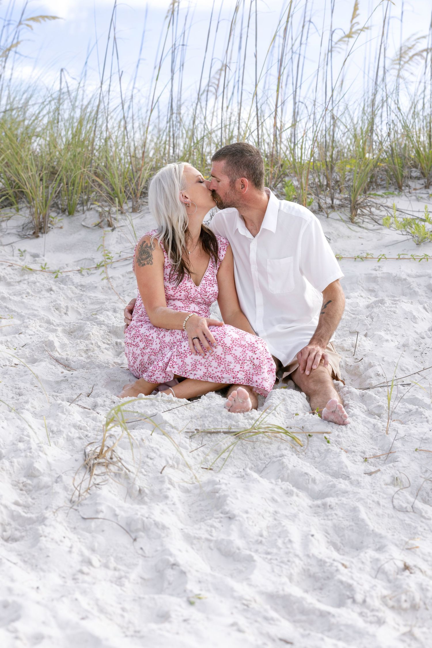 Romantic couple sharing a kiss while sitting on white sand beach dunes together.