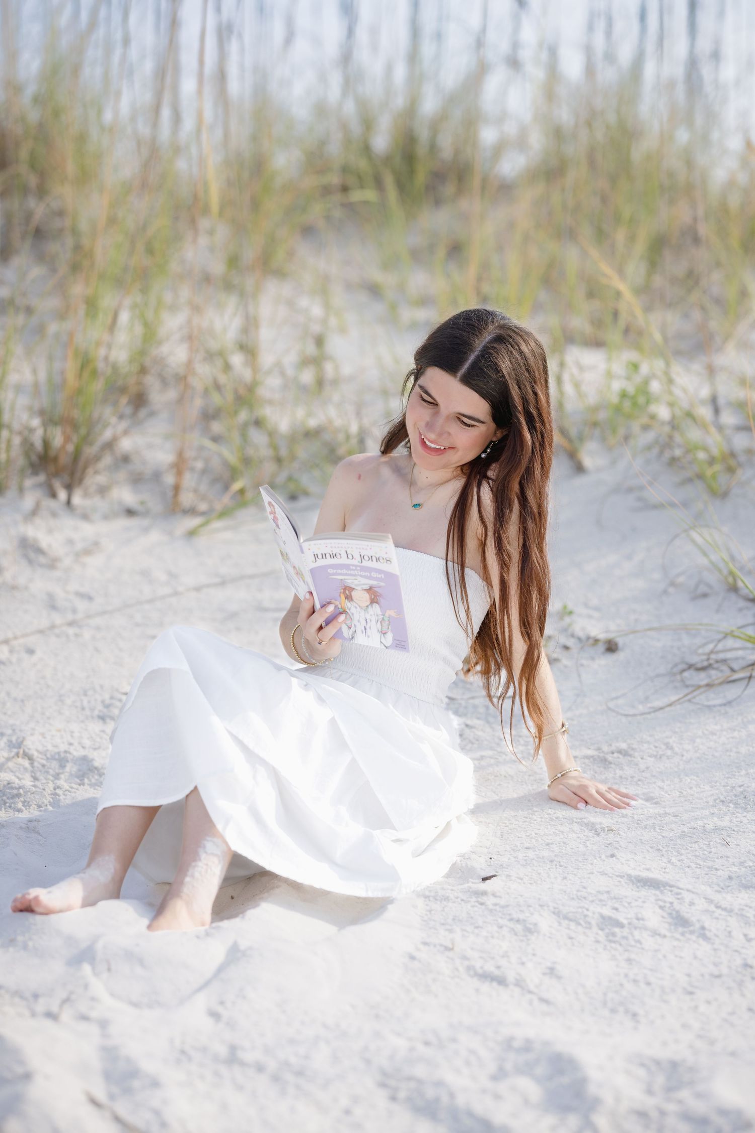 Young woman in white dress reading a book while sitting barefoot on sandy beach dunes.