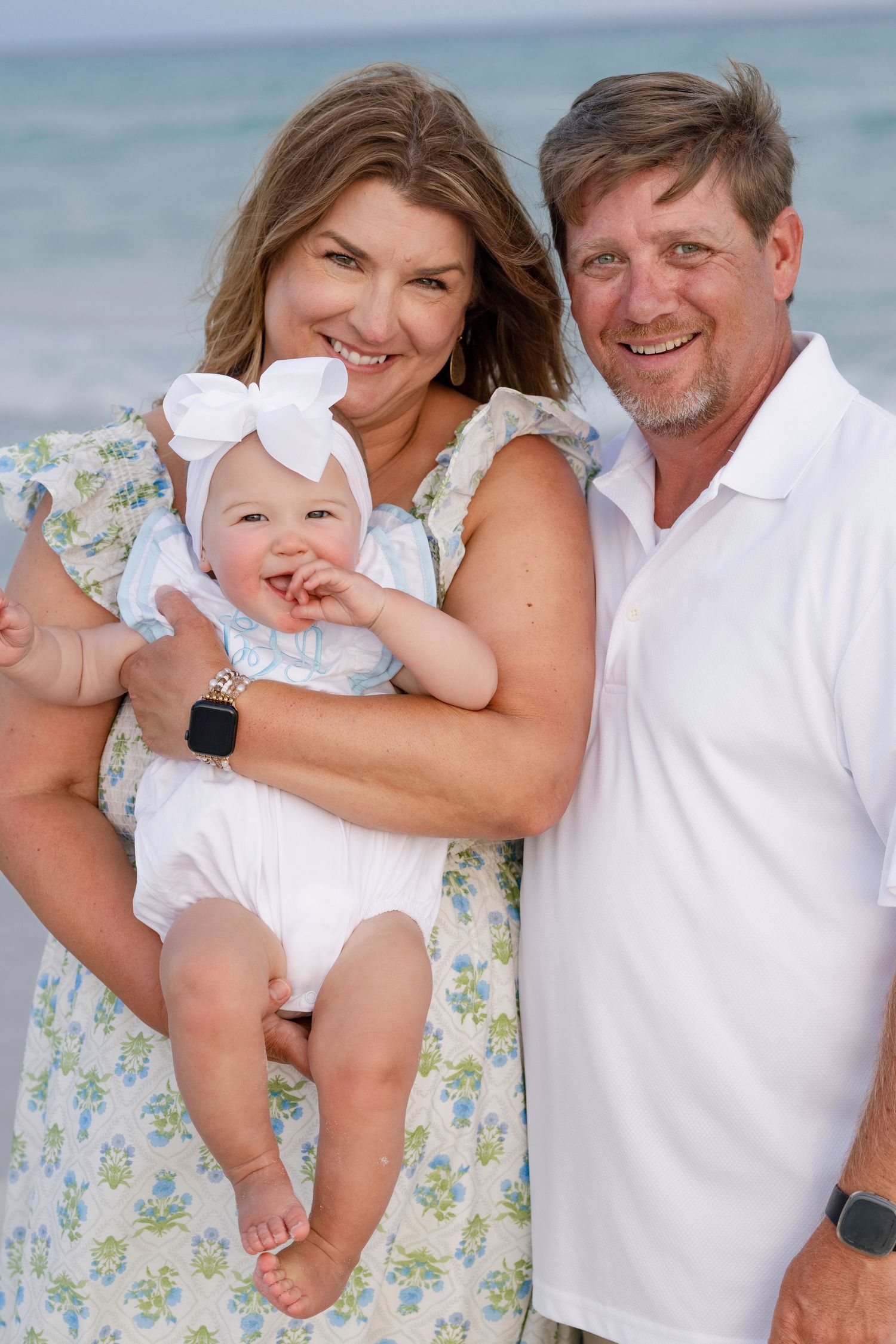 Grandparents smiling and holding adorable baby girl with bow headband at the beach.
