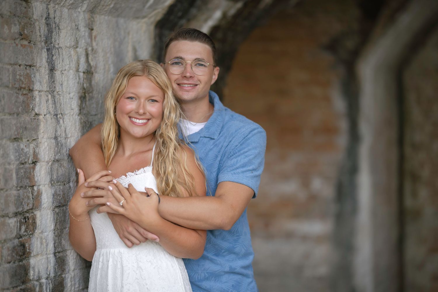 A smiling young couple poses together against a rustic brick wall, the man embracing the woman in a white dress.