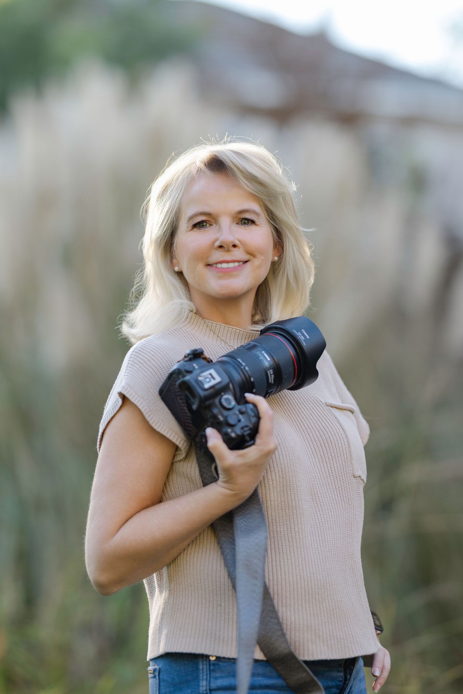 A photographer holds professional camera equipment while posing outdoors near greenery.