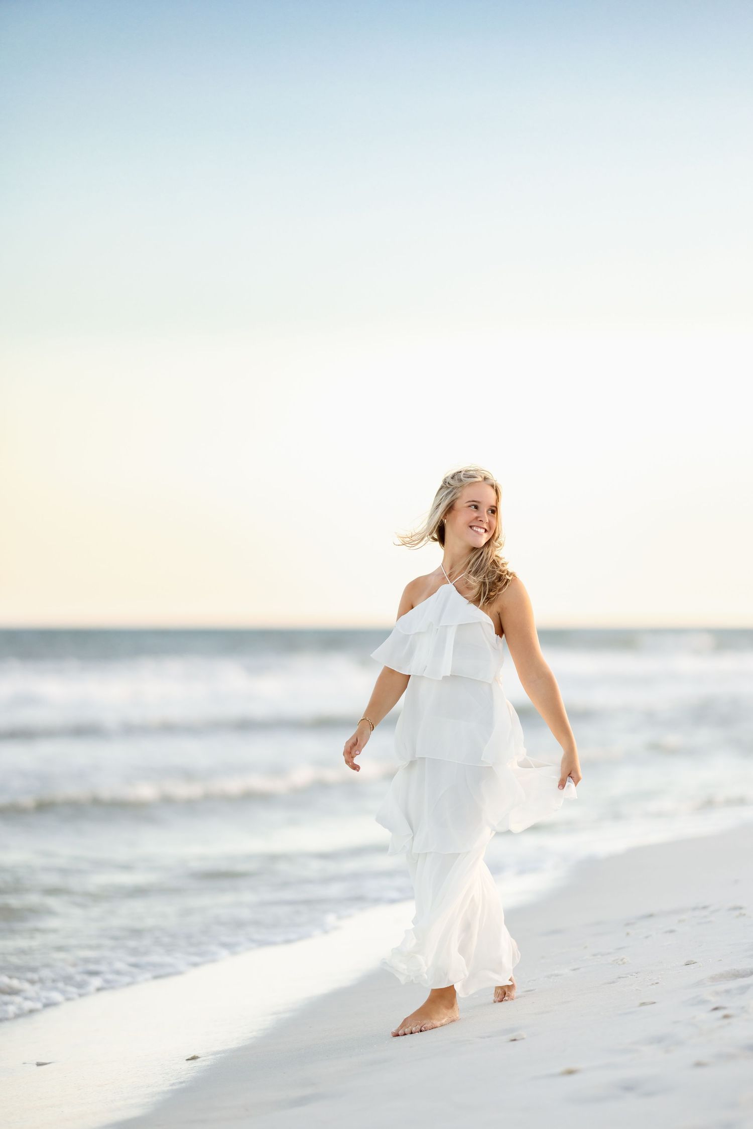 A smiling blonde woman in a white ruffled dress walks barefoot along a sandy beach at sunset.