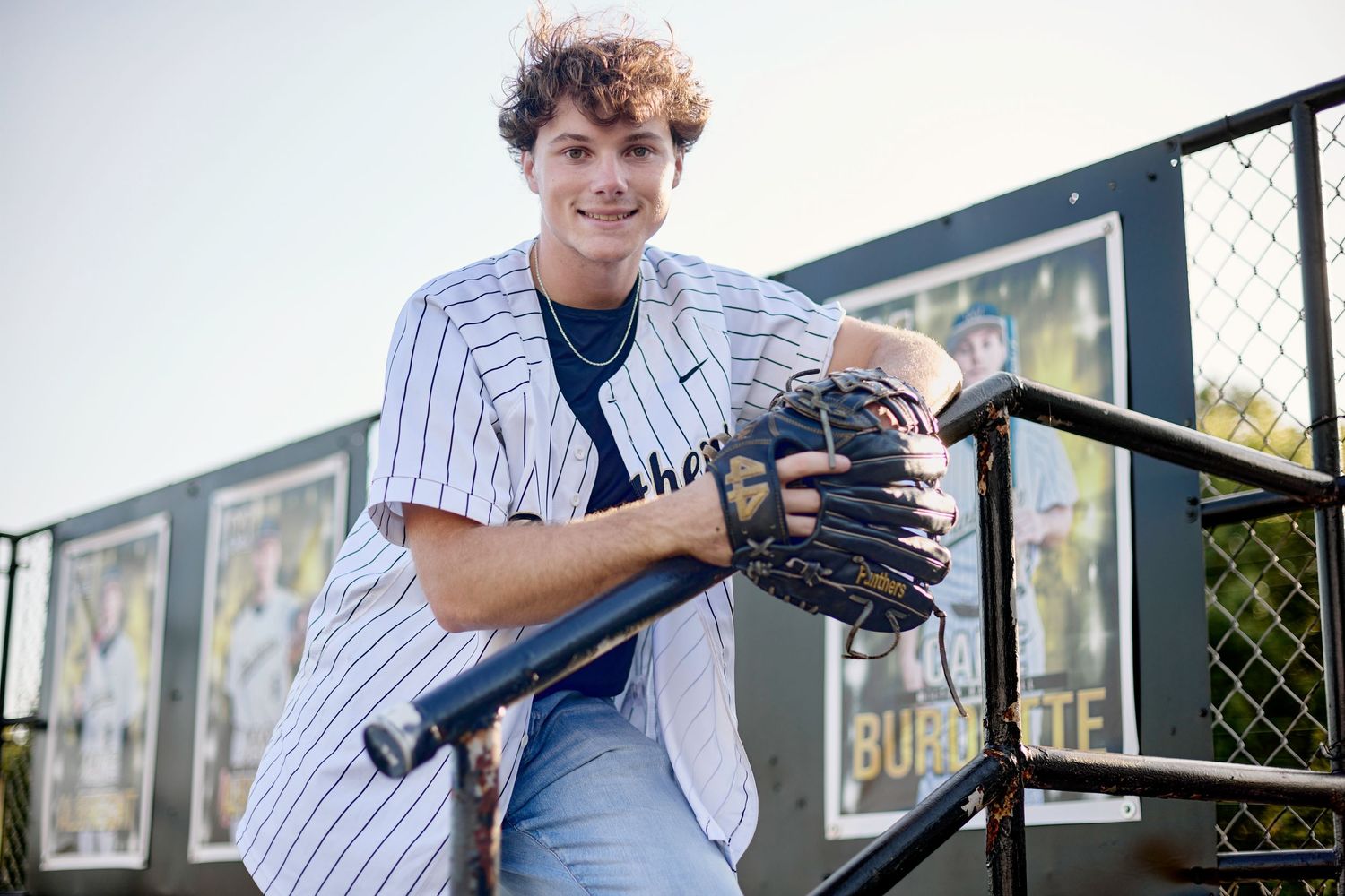 Teen baseball player in pinstripe jersey holding bat and glove at outdoor ballpark.
