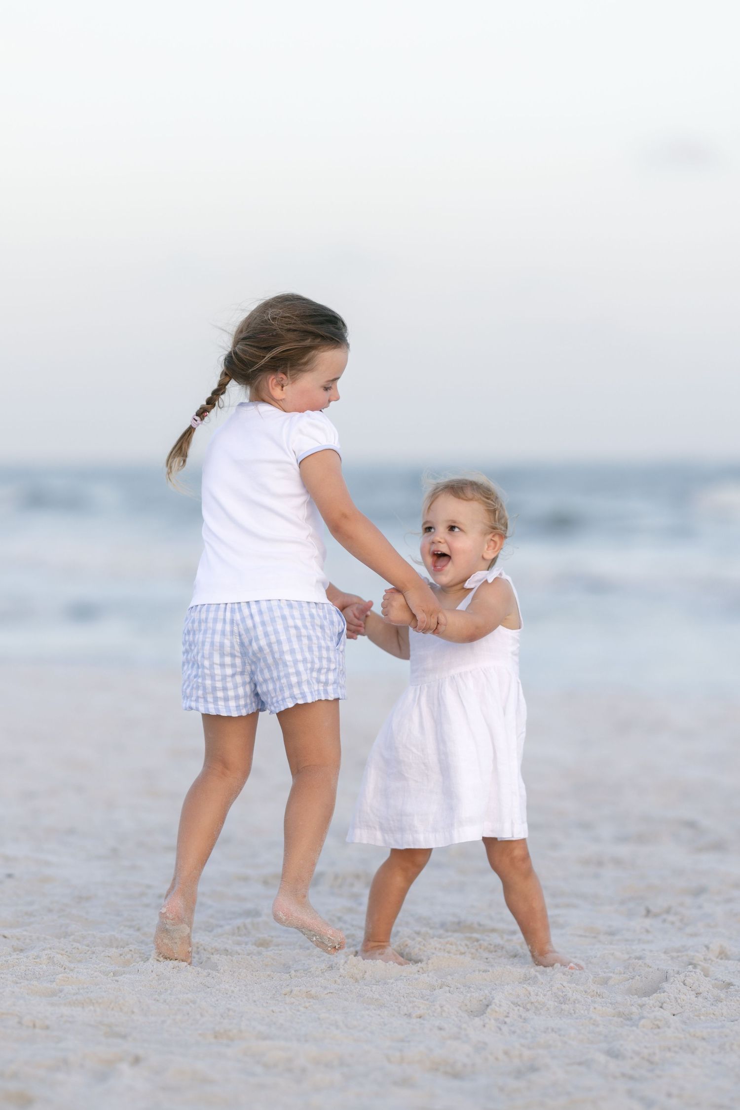 Two young sisters holding hands and playing together on a sandy beach near the ocean waves.