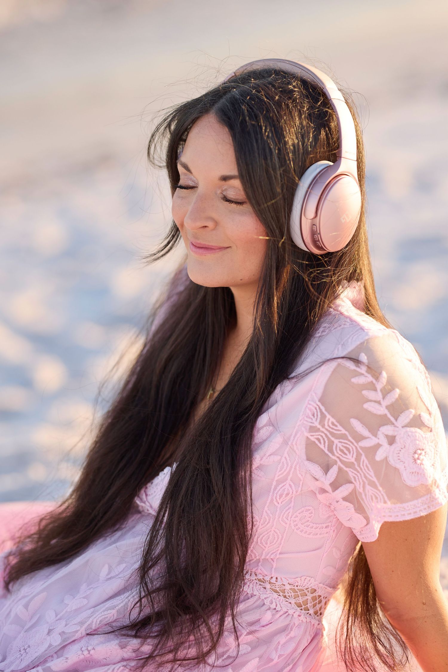 Serene woman in pink lace dress with rose gold headphones sitting on beach with eyes closed.