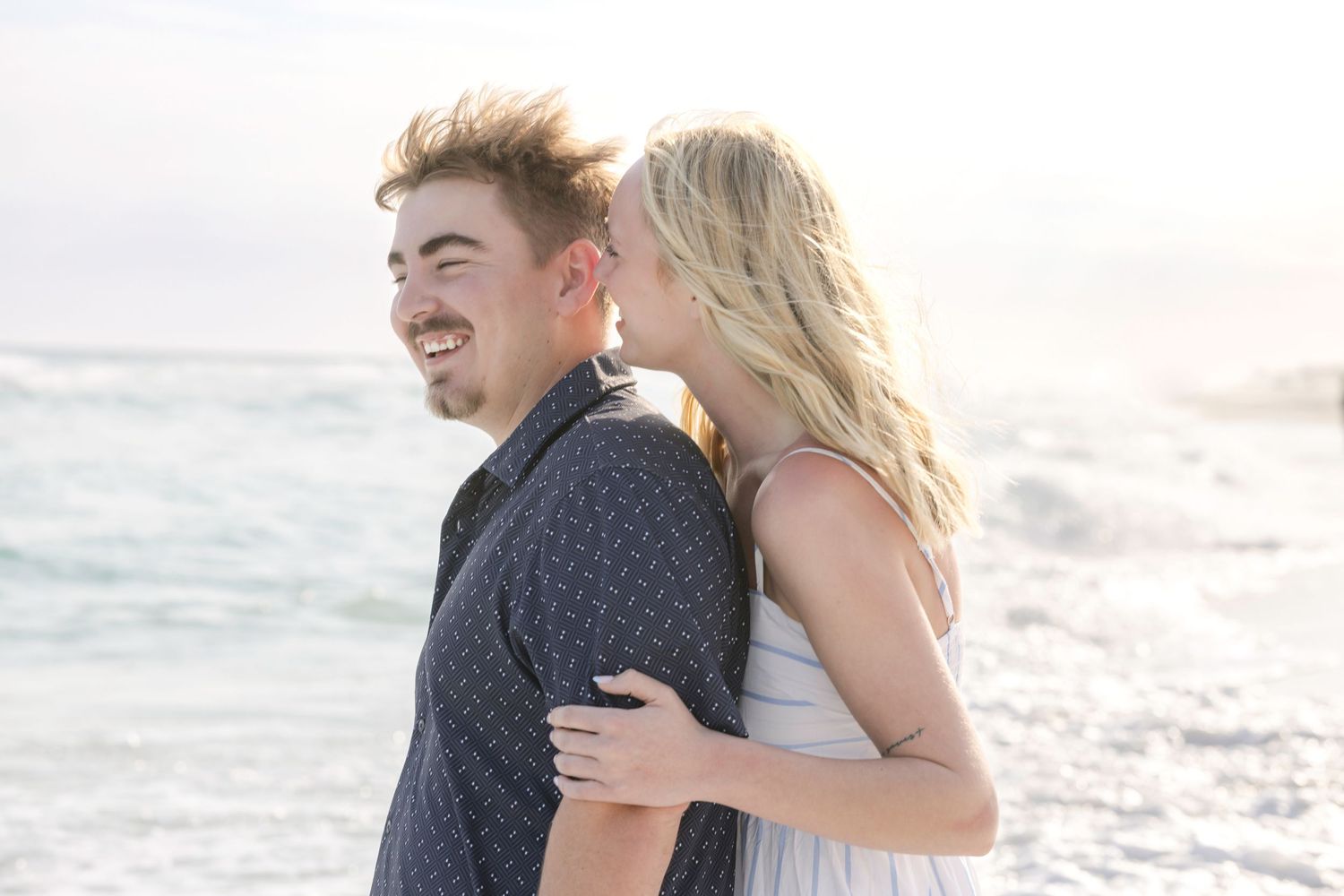 Smiling couple hugging on a bright beach with ocean waves behind them.