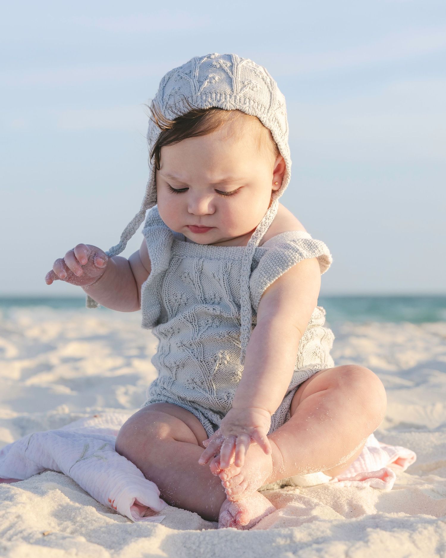Adorable baby in knit romper and bonnet sitting on white sand beach, looking down curiously.