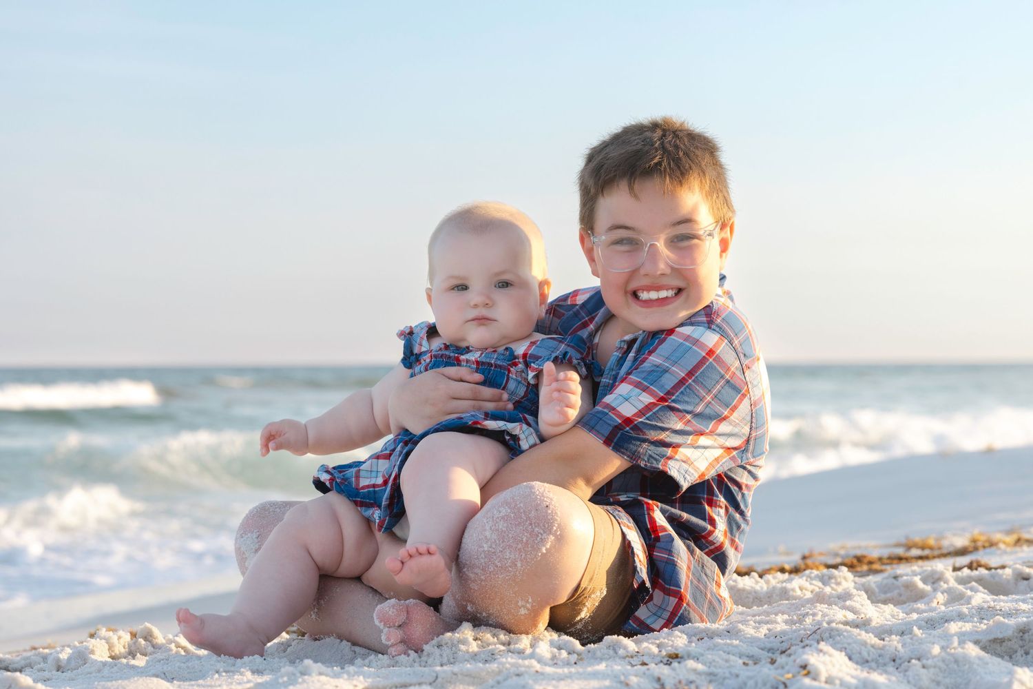 Two young boys, a toddler and older brother, smile while sitting together on a sandy beach with ocean waves behind them.