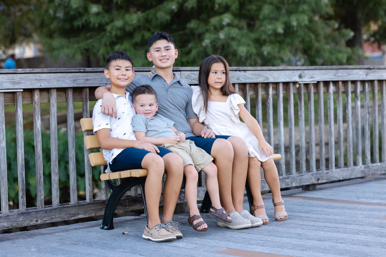 Four children sitting together on a wooden bench outdoors, smiling for a family portrait photo.