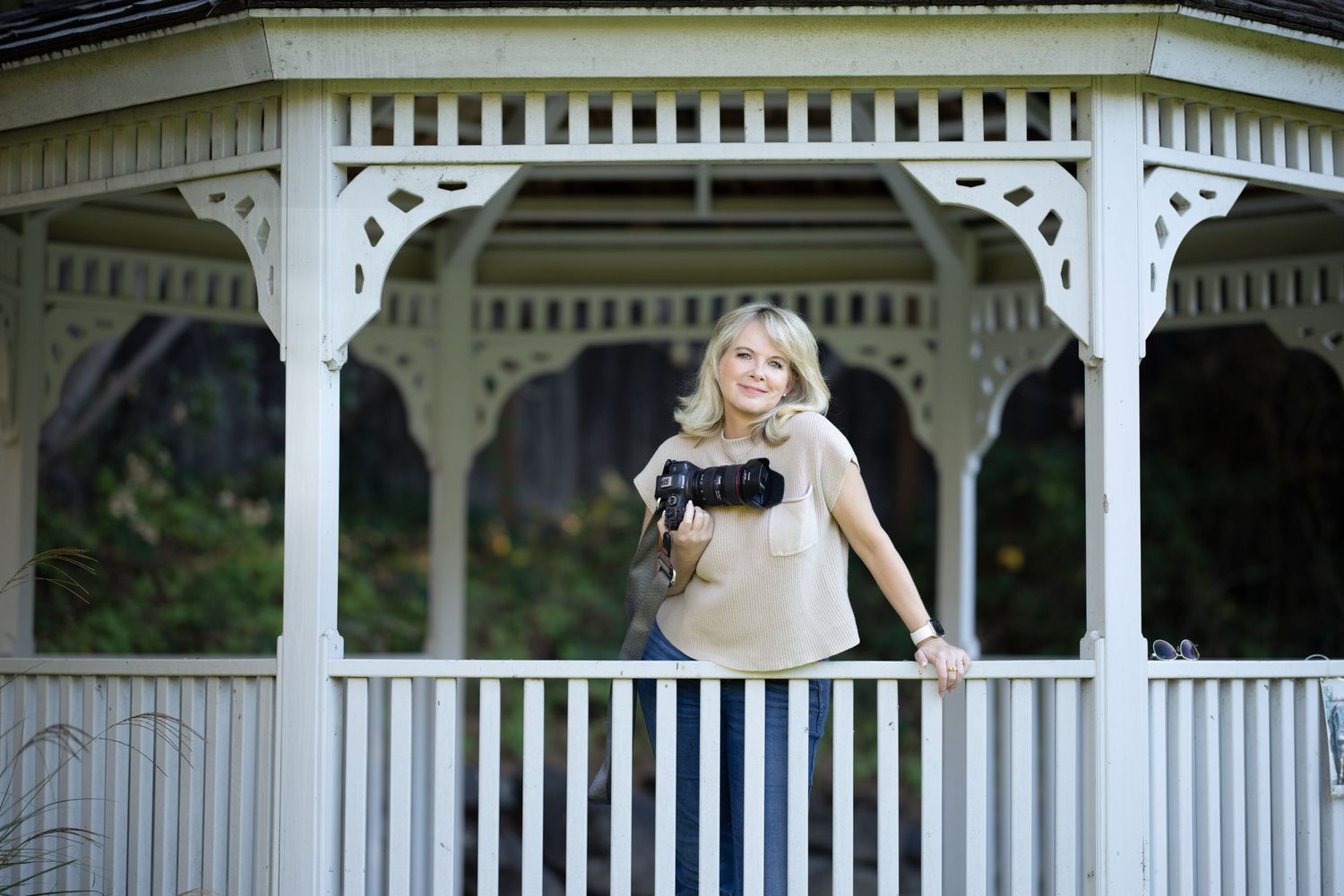A photographer with a camera poses near a white gazebo on a sunny day.