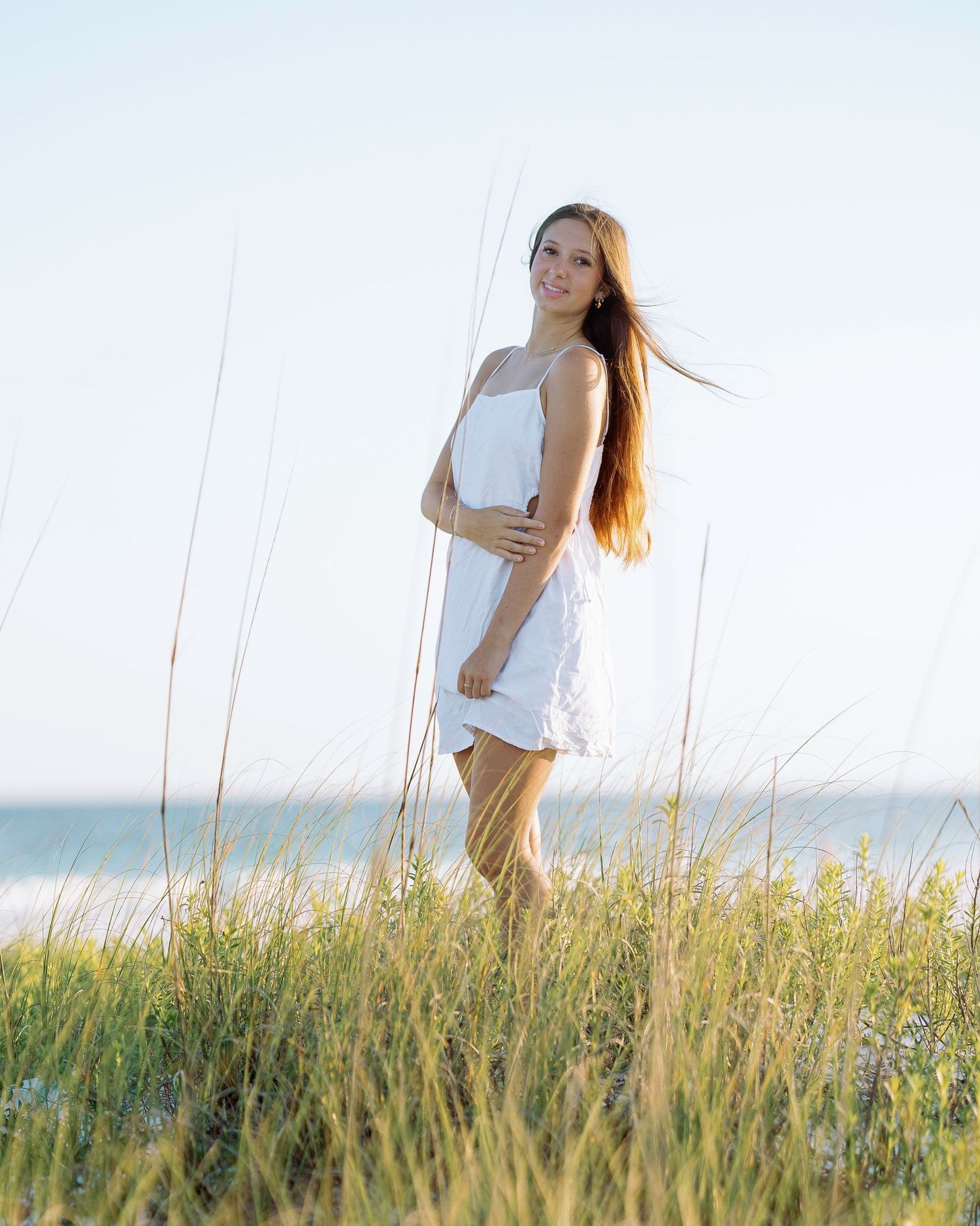 A person in white clothing stands gracefully among tall beach grass with the ocean in the background.
