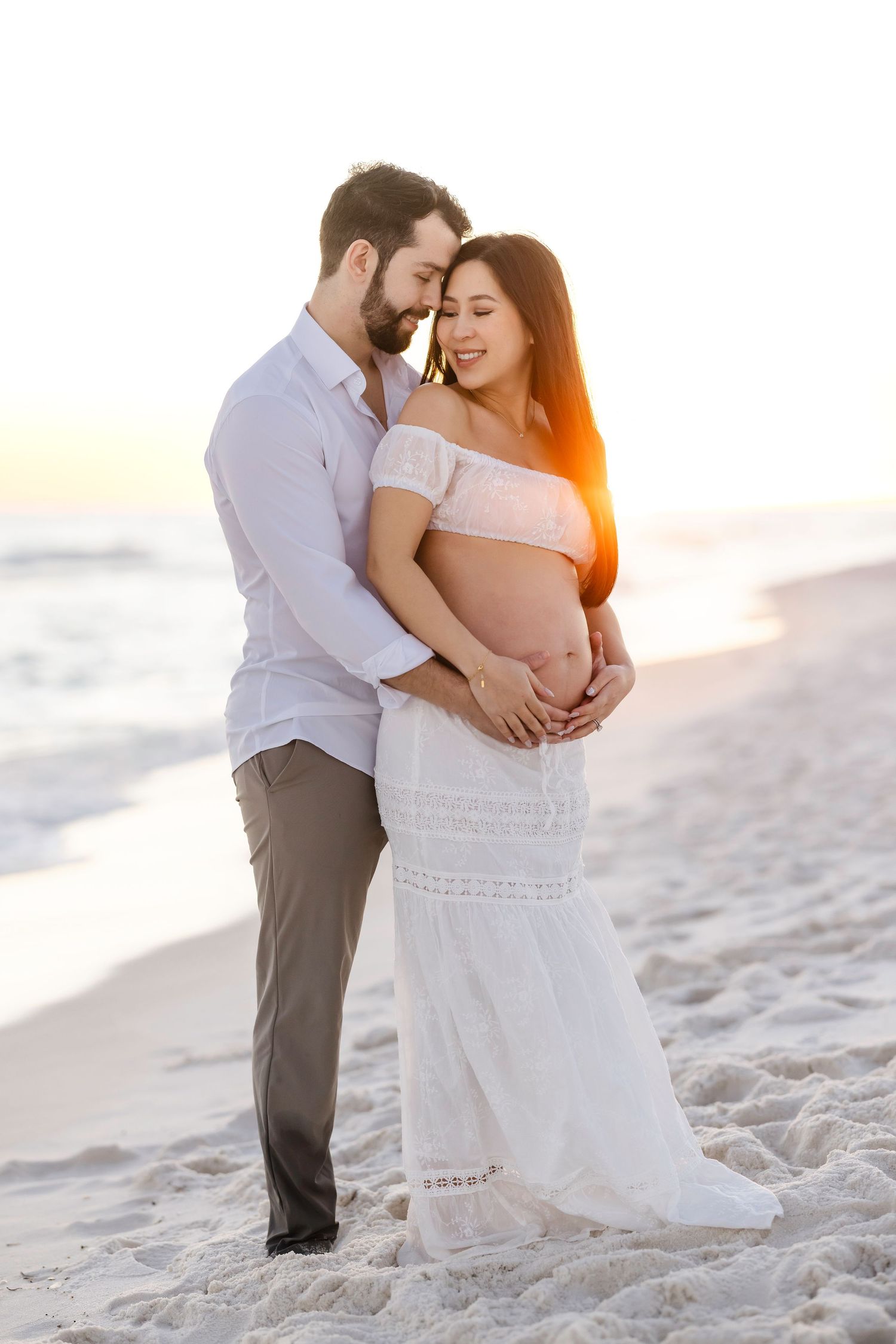 A couple embraces on a sandy beach at sunset with soft golden light illuminating their romantic moment.