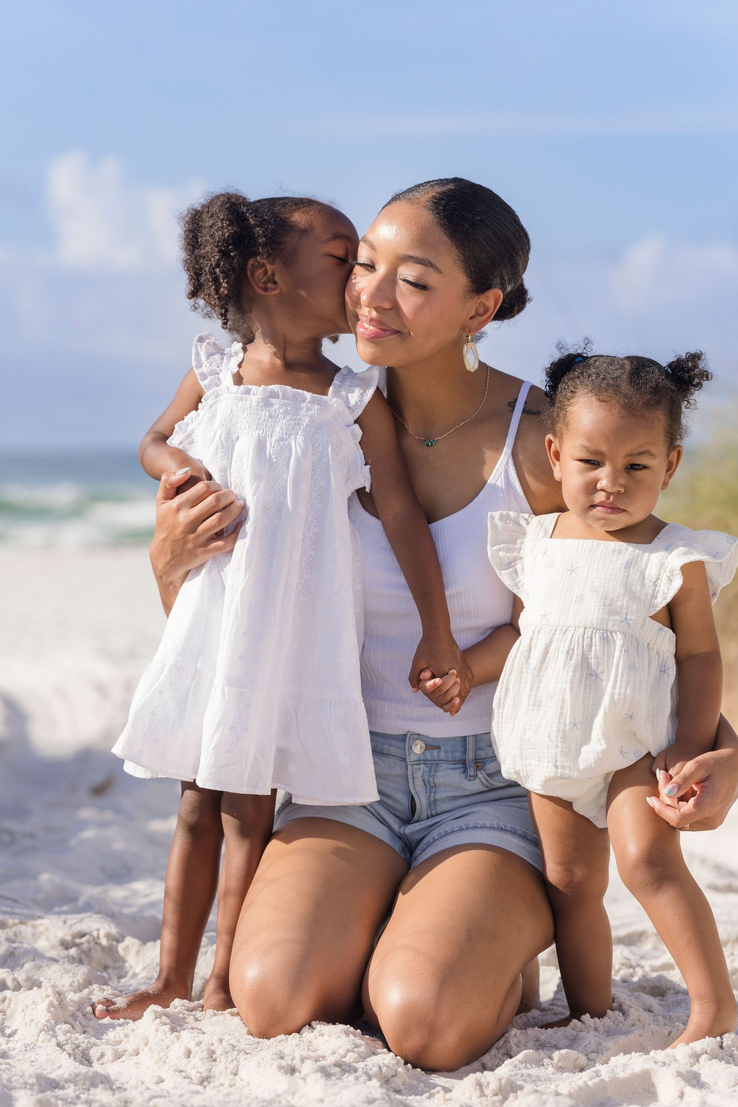 A mother kneels on white sand beach with her two young daughters in matching white dresses, sharing a tender moment.