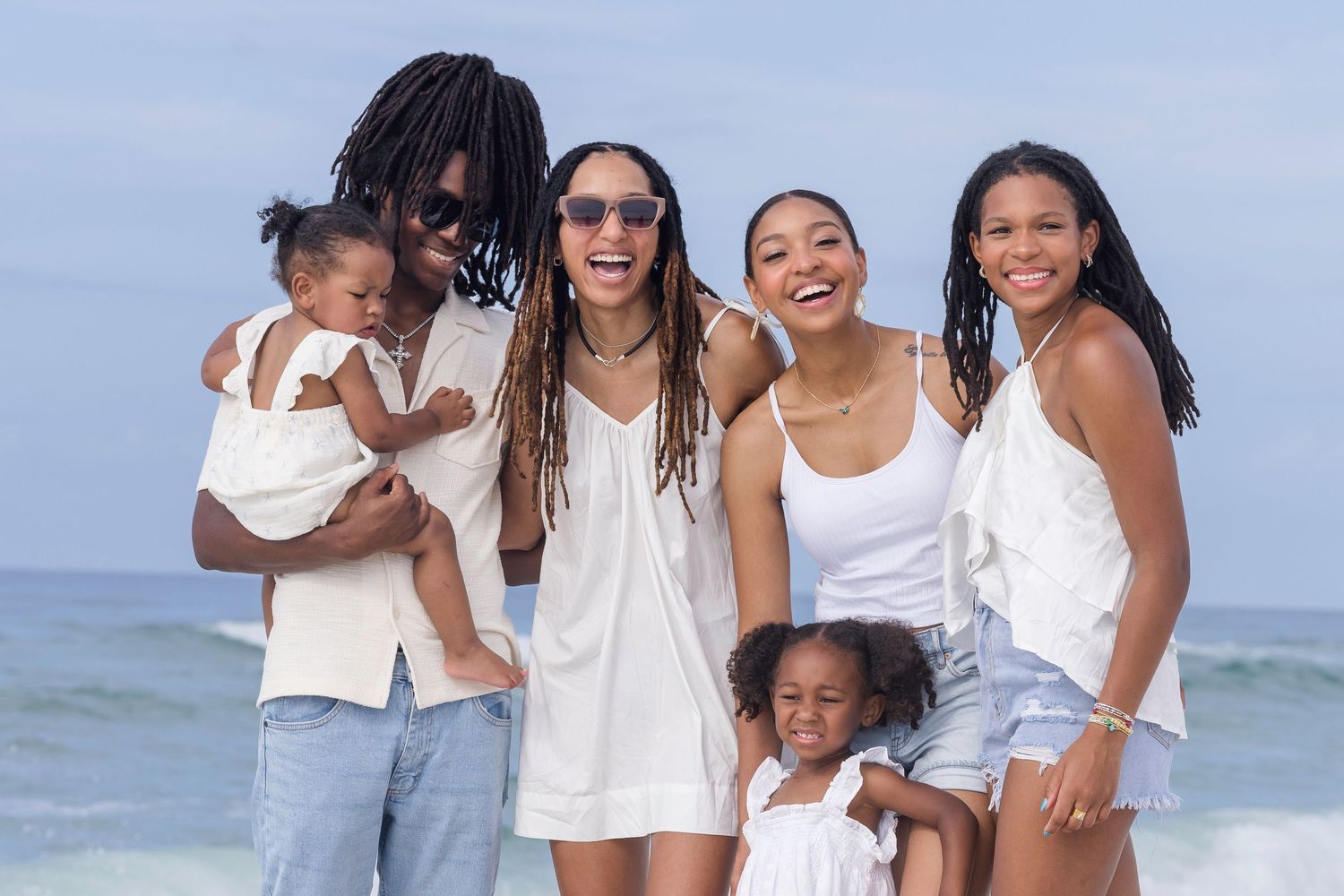 Happy family of six smiling together on a sunny beach, dressed in white outfits with ocean waves in the background.