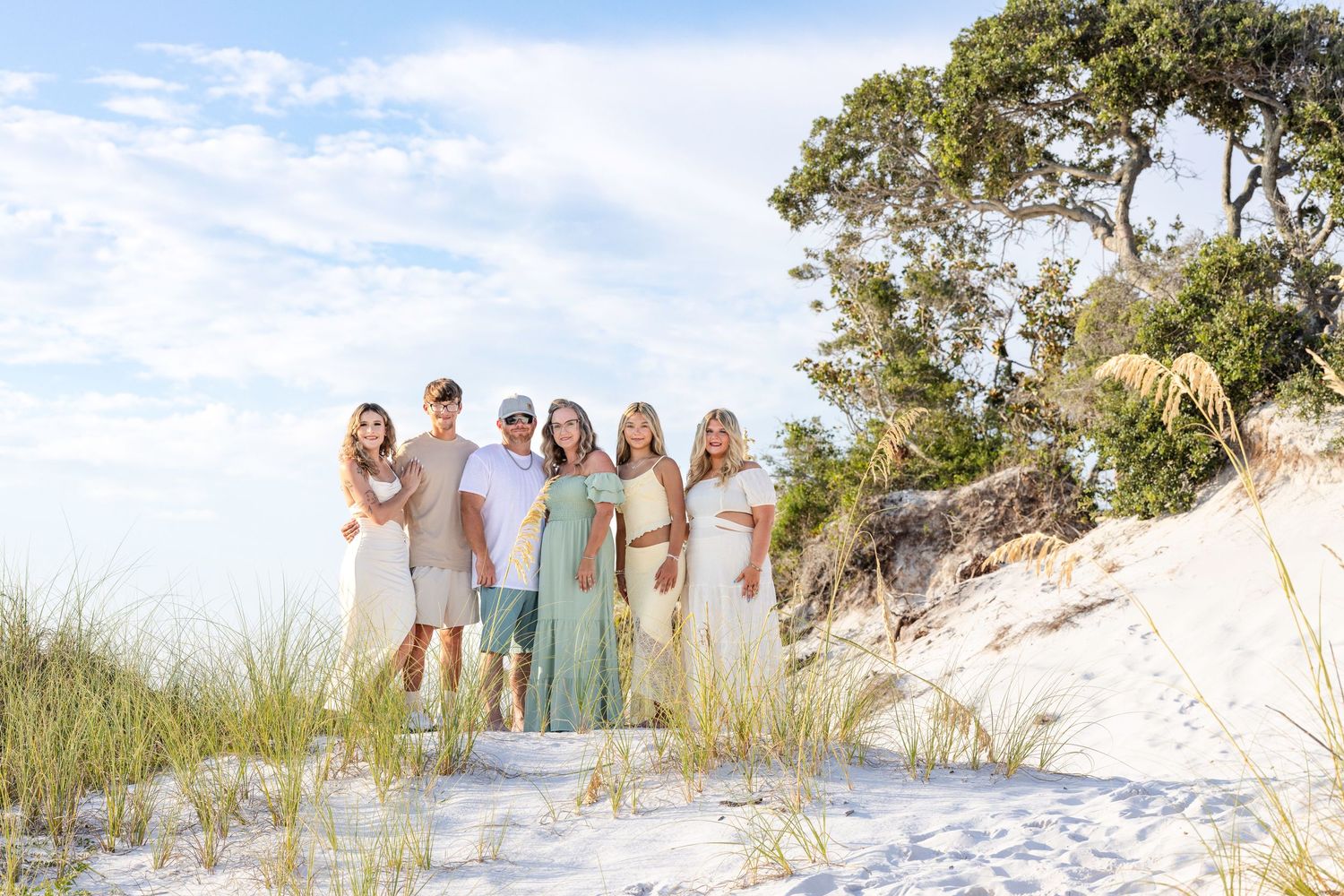 Family of seven posing together on white sand beach dunes with lush green trees and blue sky backdrop.