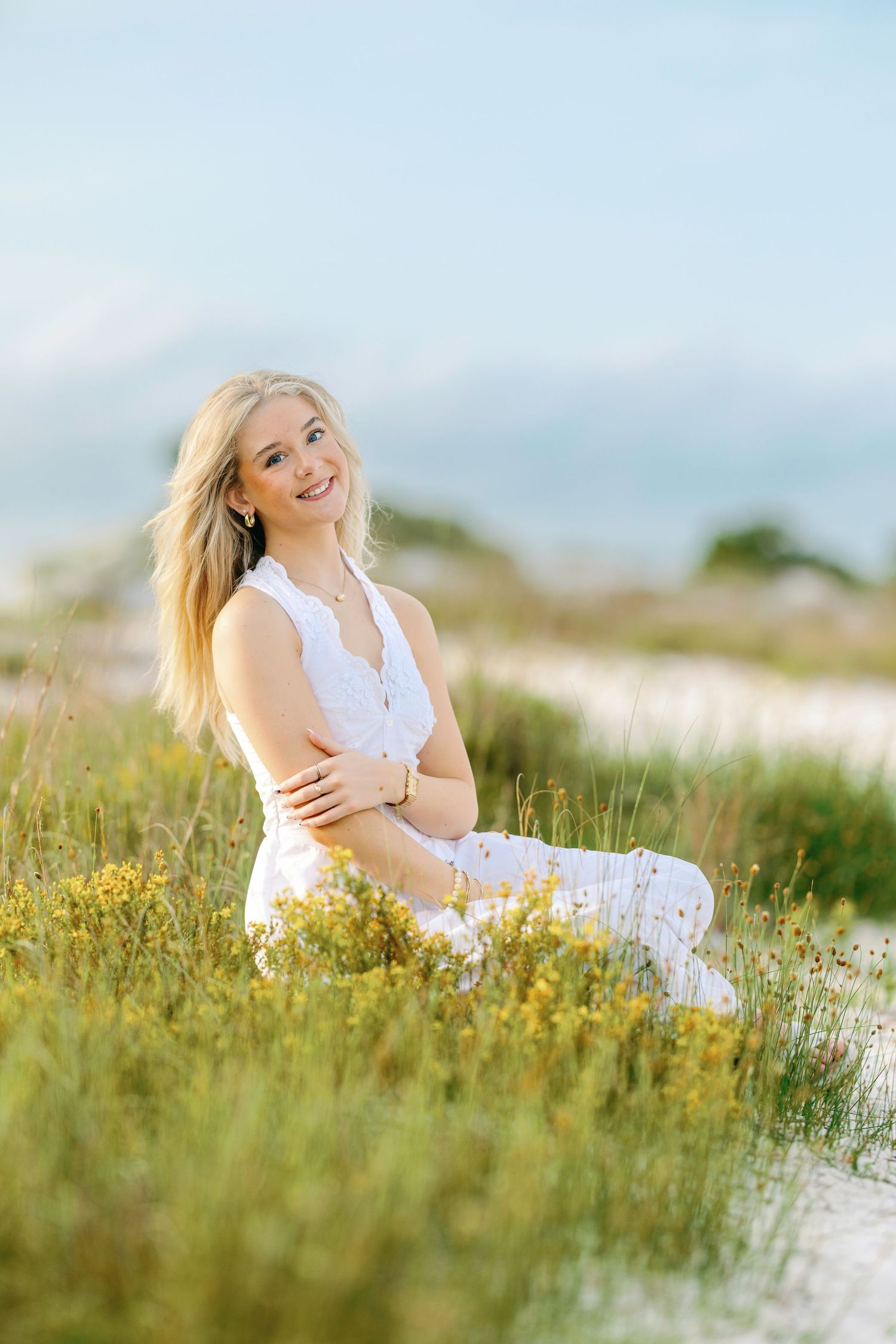 Smiling blonde woman in white outfit sitting among wildflowers on sandy dunes with mountains in background.