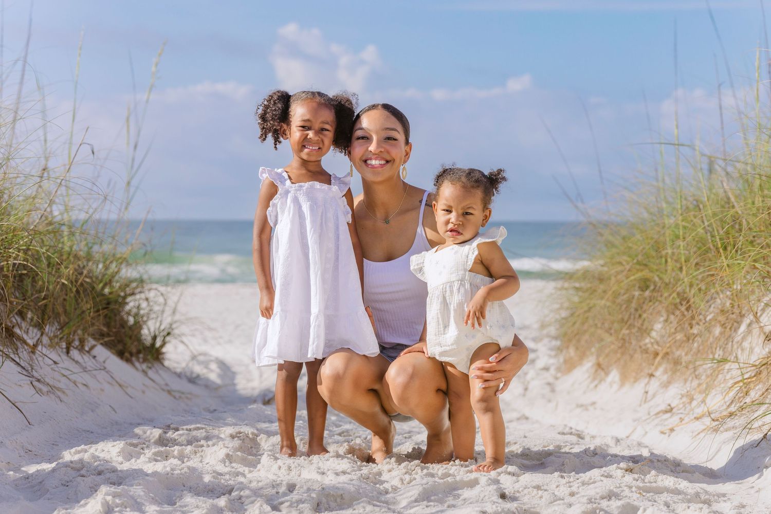 A smiling mother crouches with her two young daughters on a sunny white sand beach near sea grass.