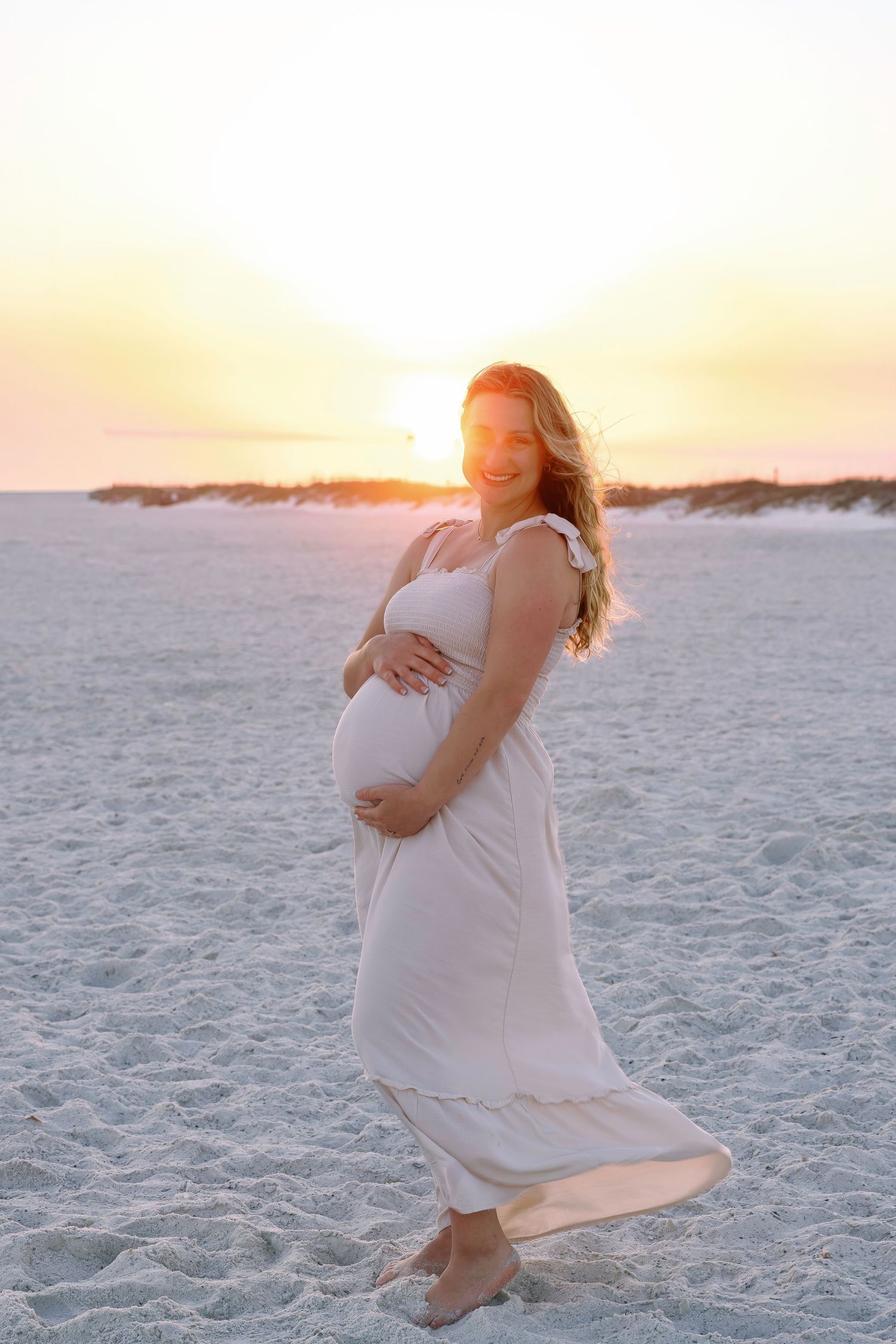 Pregnant woman in white dress cradling baby bump on sandy beach at golden sunset.