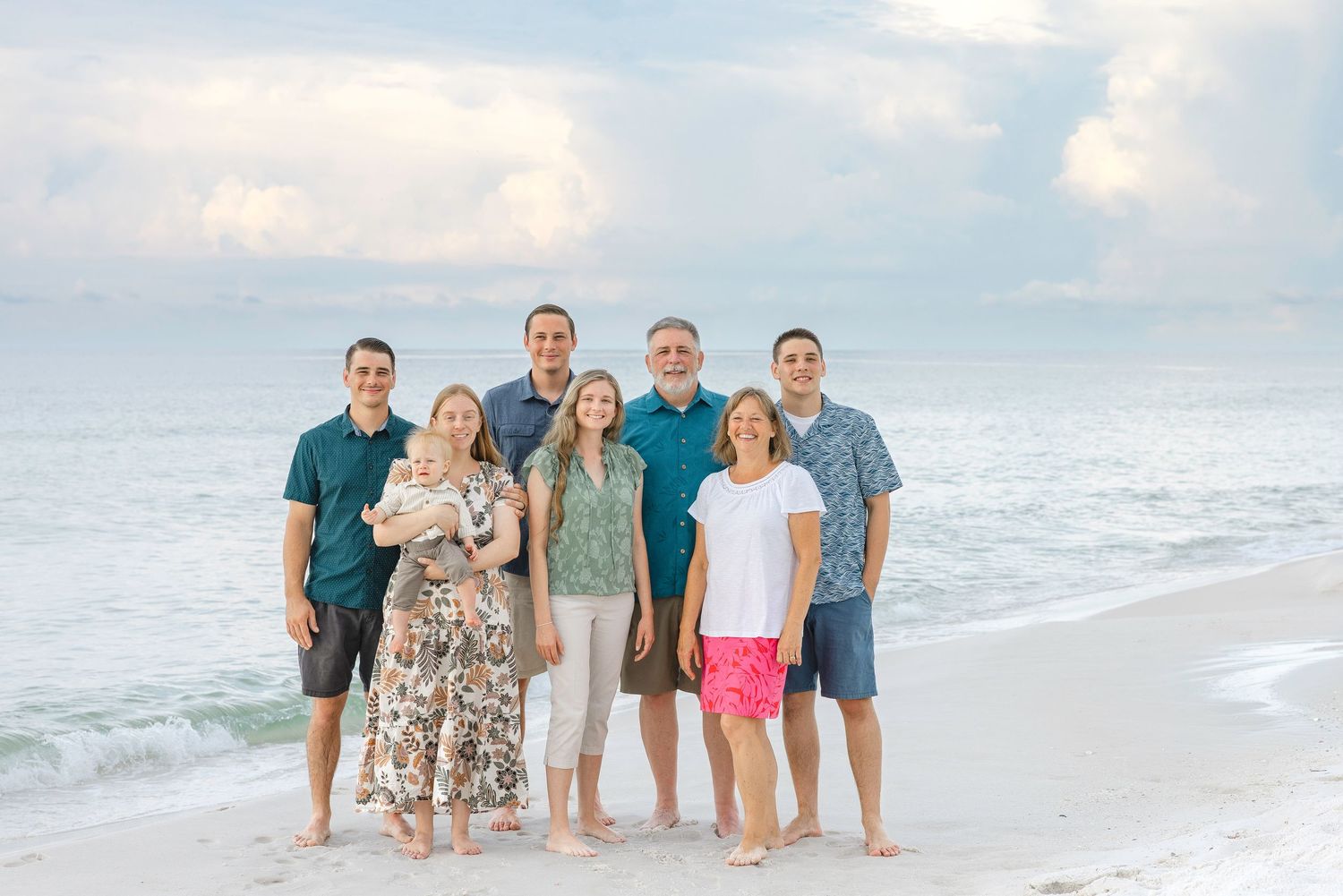 Family group wearing coordinated blue and floral outfits posing together on sunny beach at sunset.
