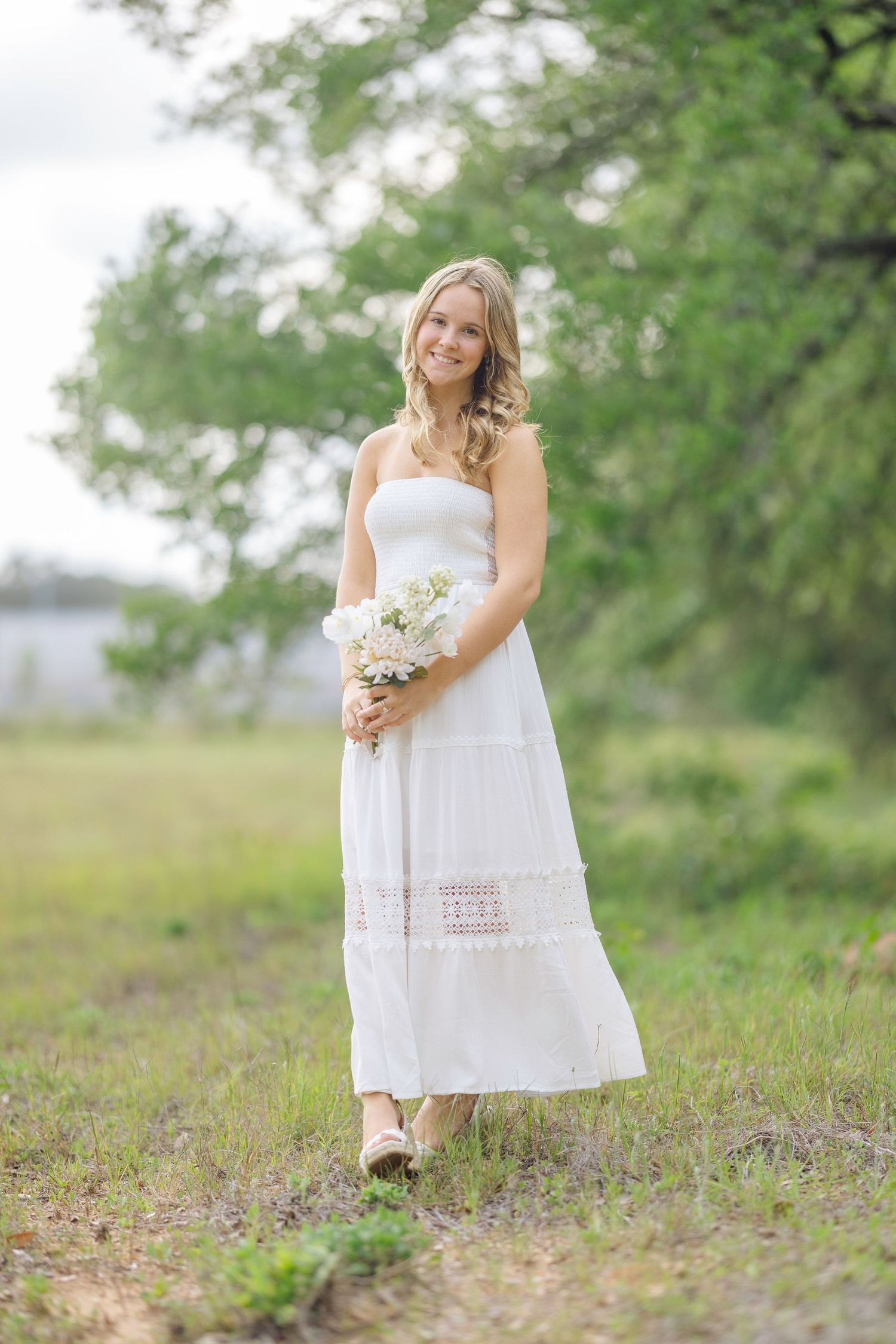 A woman in a flowing white sundress holds a bouquet of white flowers while standing in a grassy outdoor setting.