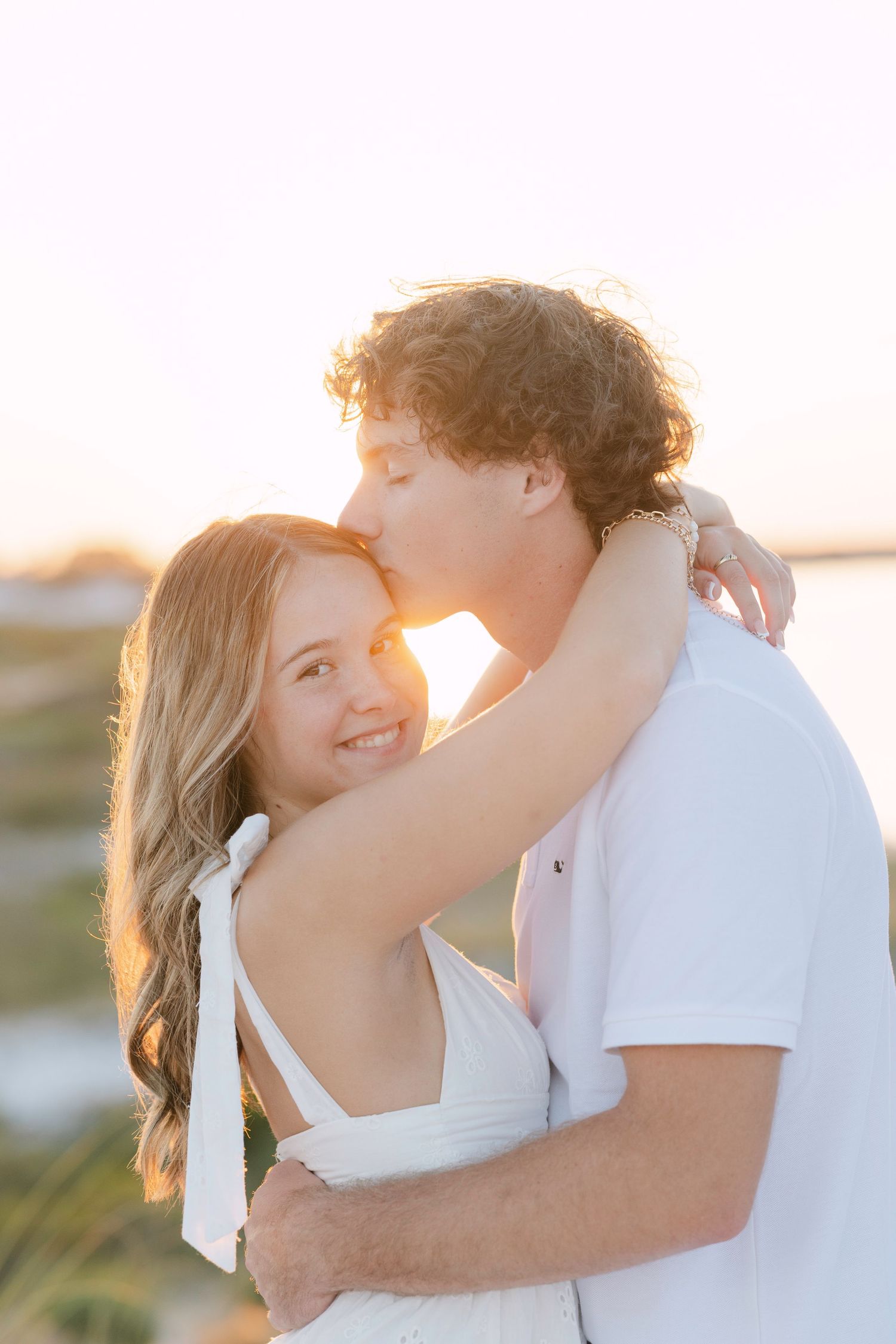 A romantic couple embraces in golden sunlight during a sunset photoshoot, wearing white outfits against a soft backlit glow.