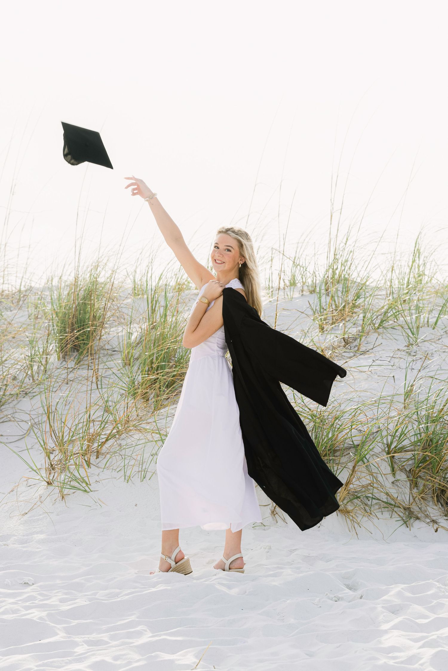 A graduate in a white dress celebrates on a sandy beach while holding a black graduation gown in various poses.