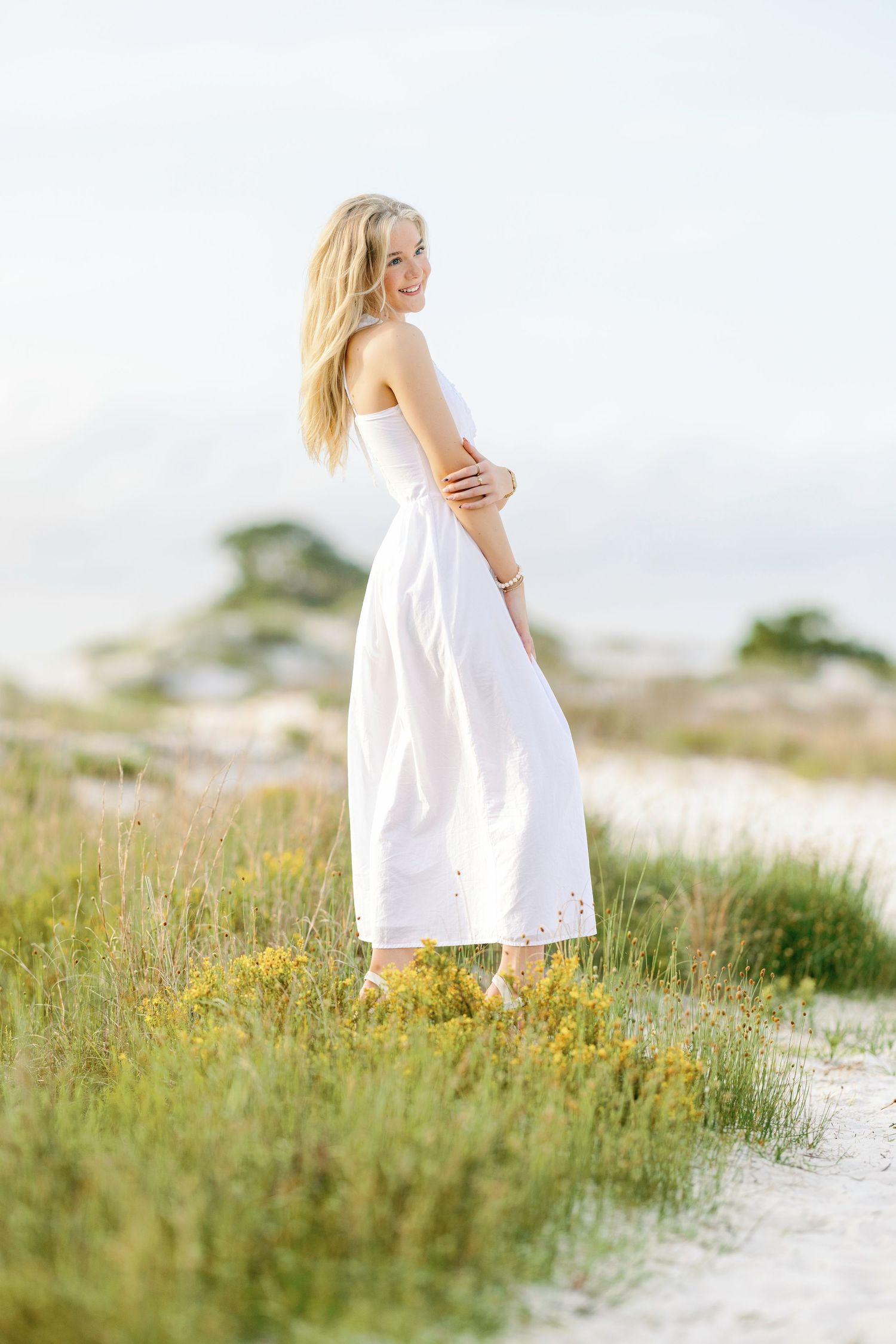Ethereal white dress flows in the coastal breeze among yellow wildflowers and dunes.