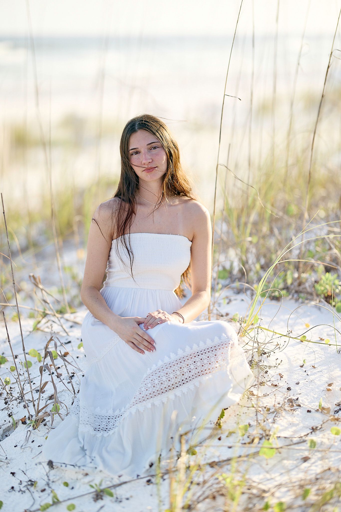 A white lace sundress stands out against beach dunes and sea grass in soft golden light.