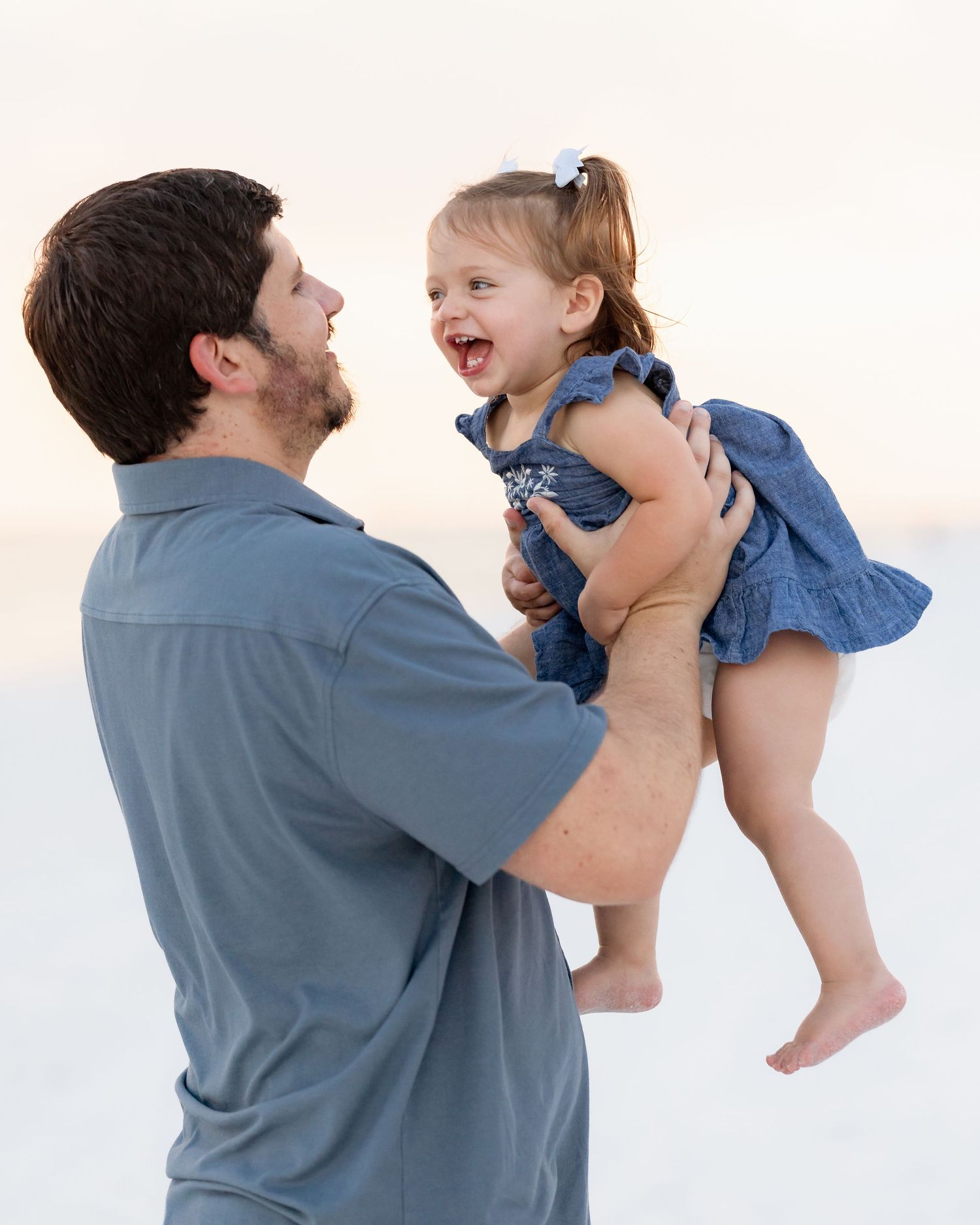 Laughing toddler girl in blue dress lifted joyfully by bearded father on bright white sand beach.